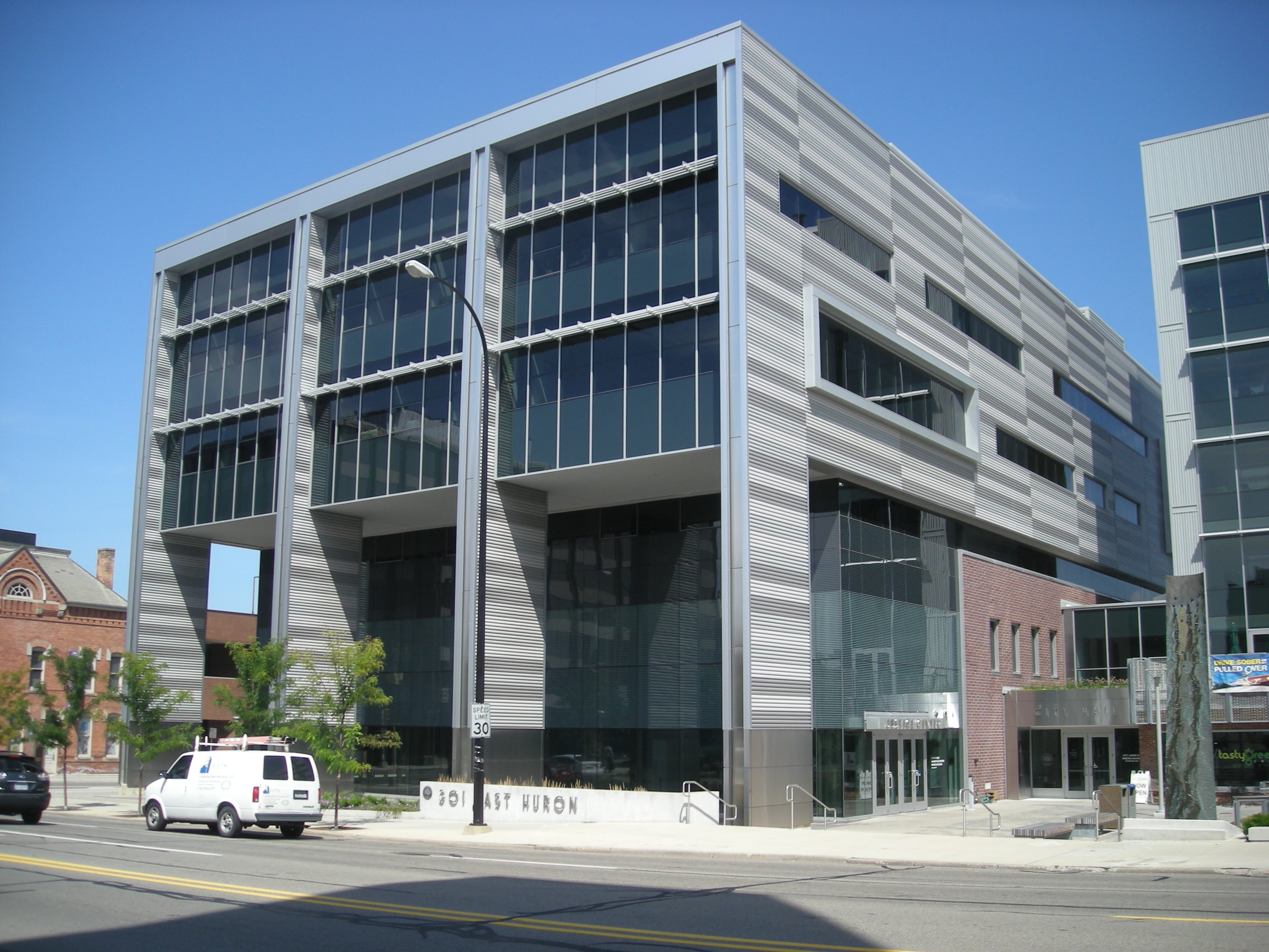 Modern glass and metal-paneled municipal building on a city street corner under blue sky.