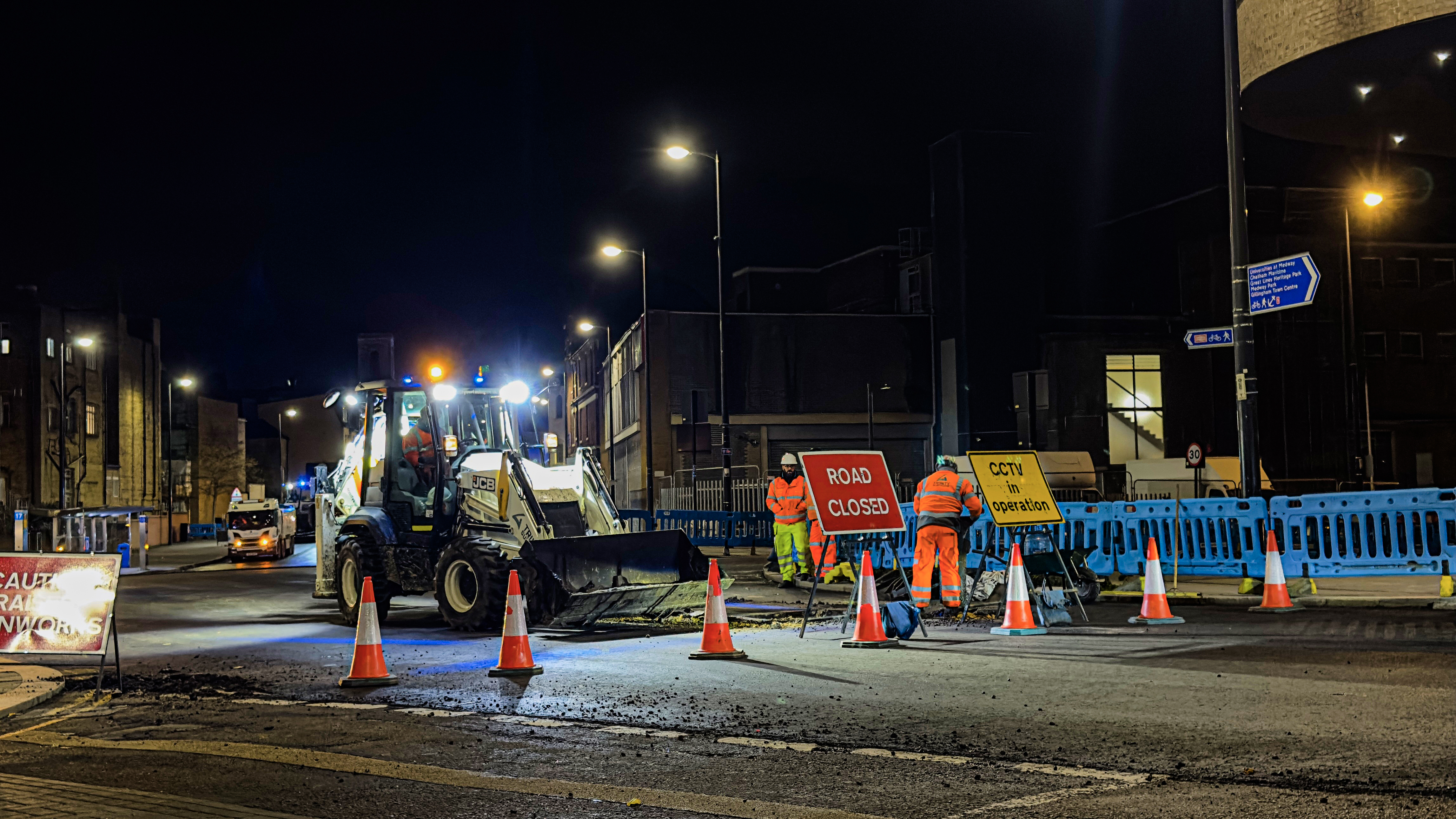 Workers and a loader at a nighttime road construction site with orange cones and a Road Closed sign.