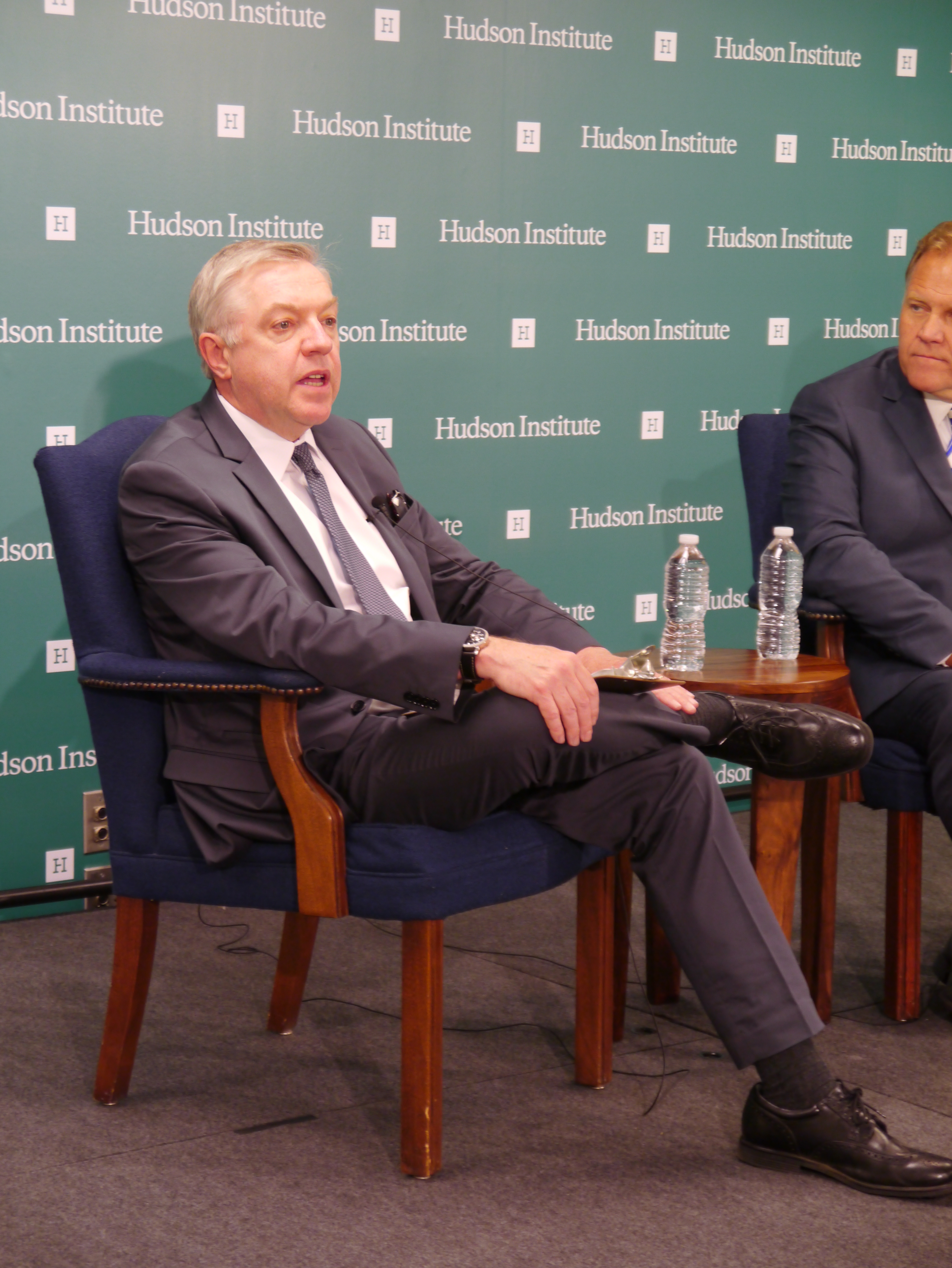 Two men in suits seated at a panel discussion in front of a branded backdrop.