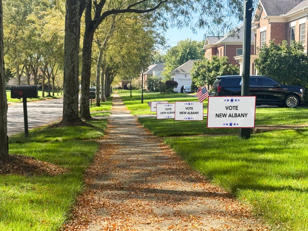 White yard signs with blue text planted in a grass strip along a suburban sidewalk.