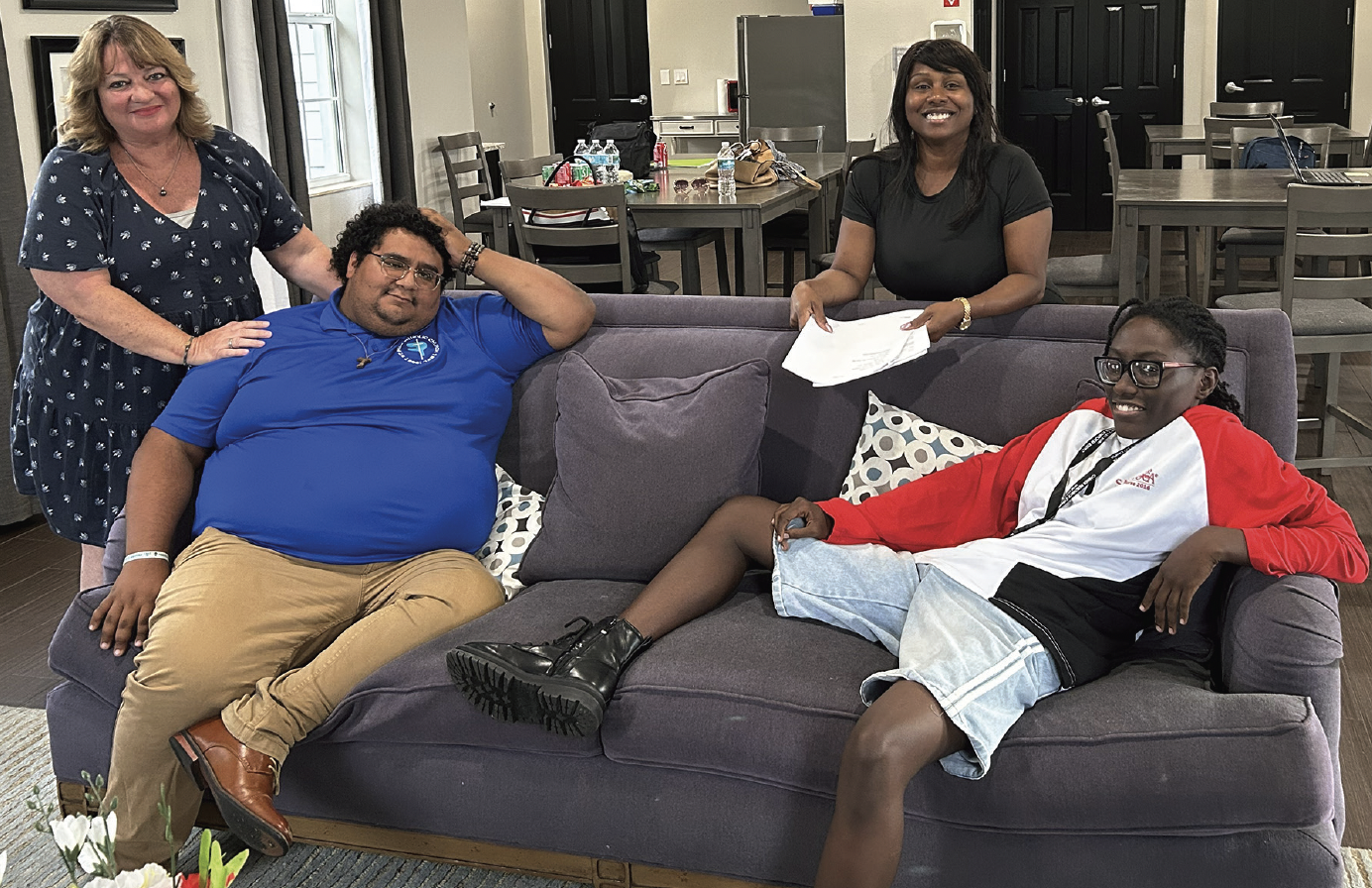 Four people in a living-room setting, two seated on a gray sofa and two standing nearby.