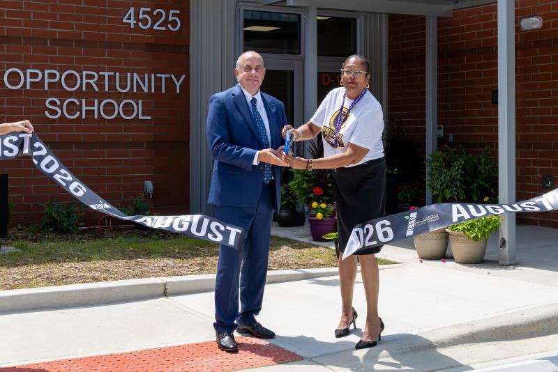 Two people cut a ribbon in front of a brick building labeled Opportunity School.