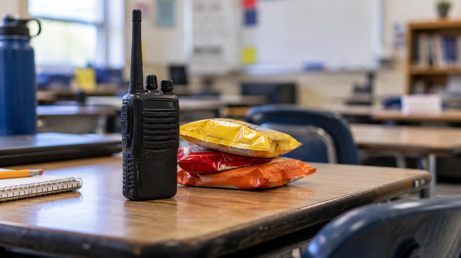 A two-way radio handset resting on a school desk beside a small stack of snack bags.