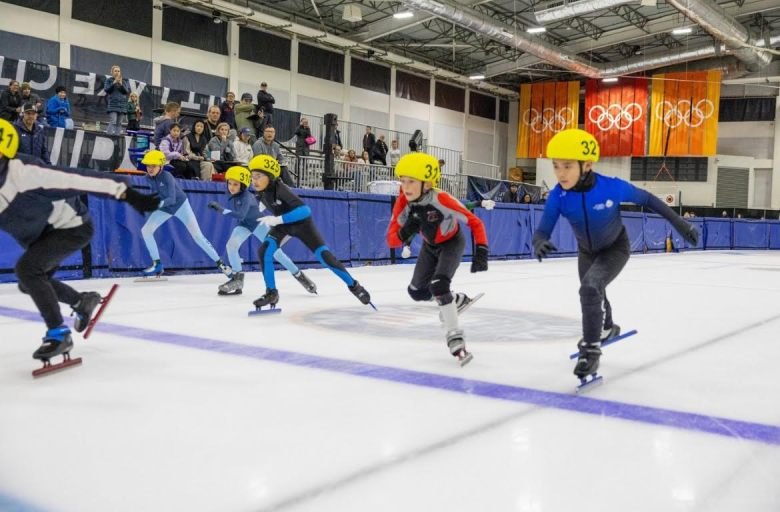 Young speedskaters in helmets and numbered bibs race on an indoor ice rink.