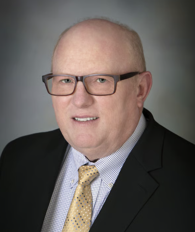 Terence Collins wears glasses, a dark jacket, a checked shirt and a yellow patterned tie against a gray background.