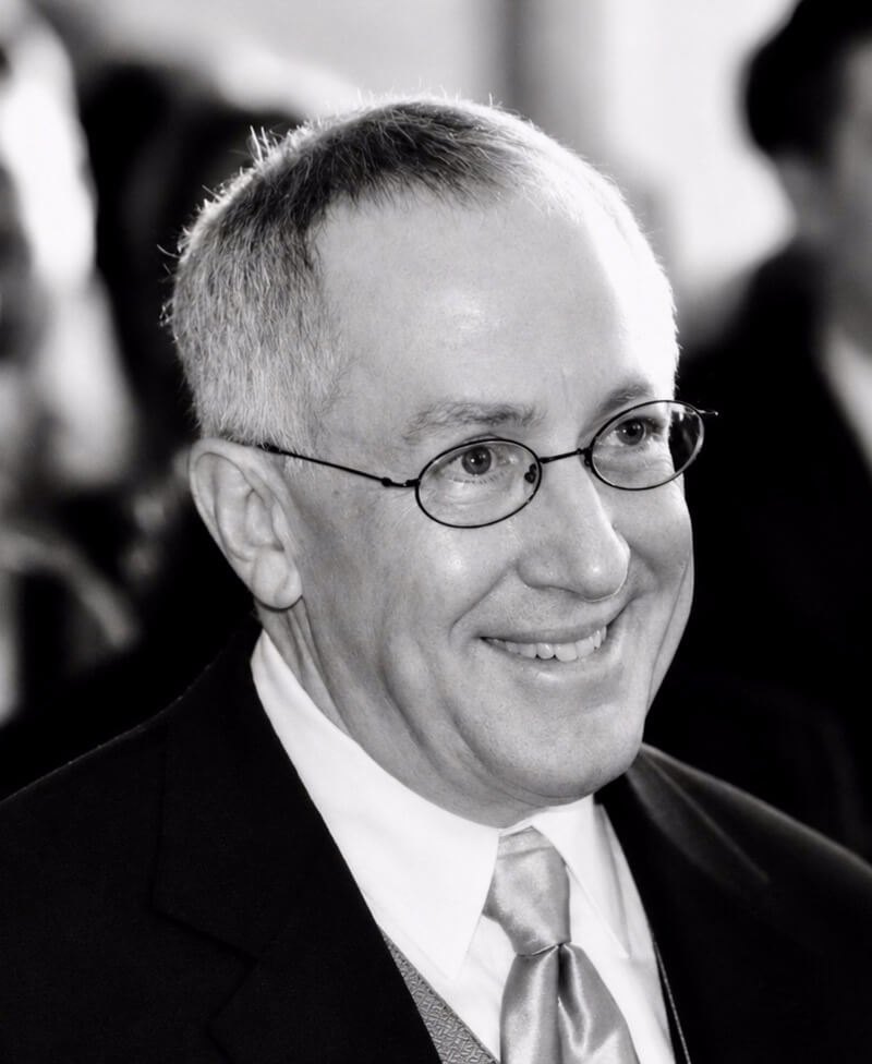 Black-and-white portrait of a smiling man in glasses, a dark suit, and a light-colored tie.
