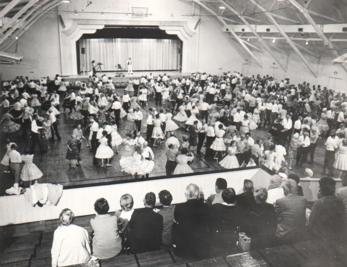 Interior of a large auditorium with exposed roof trusses and dancers on the floor.