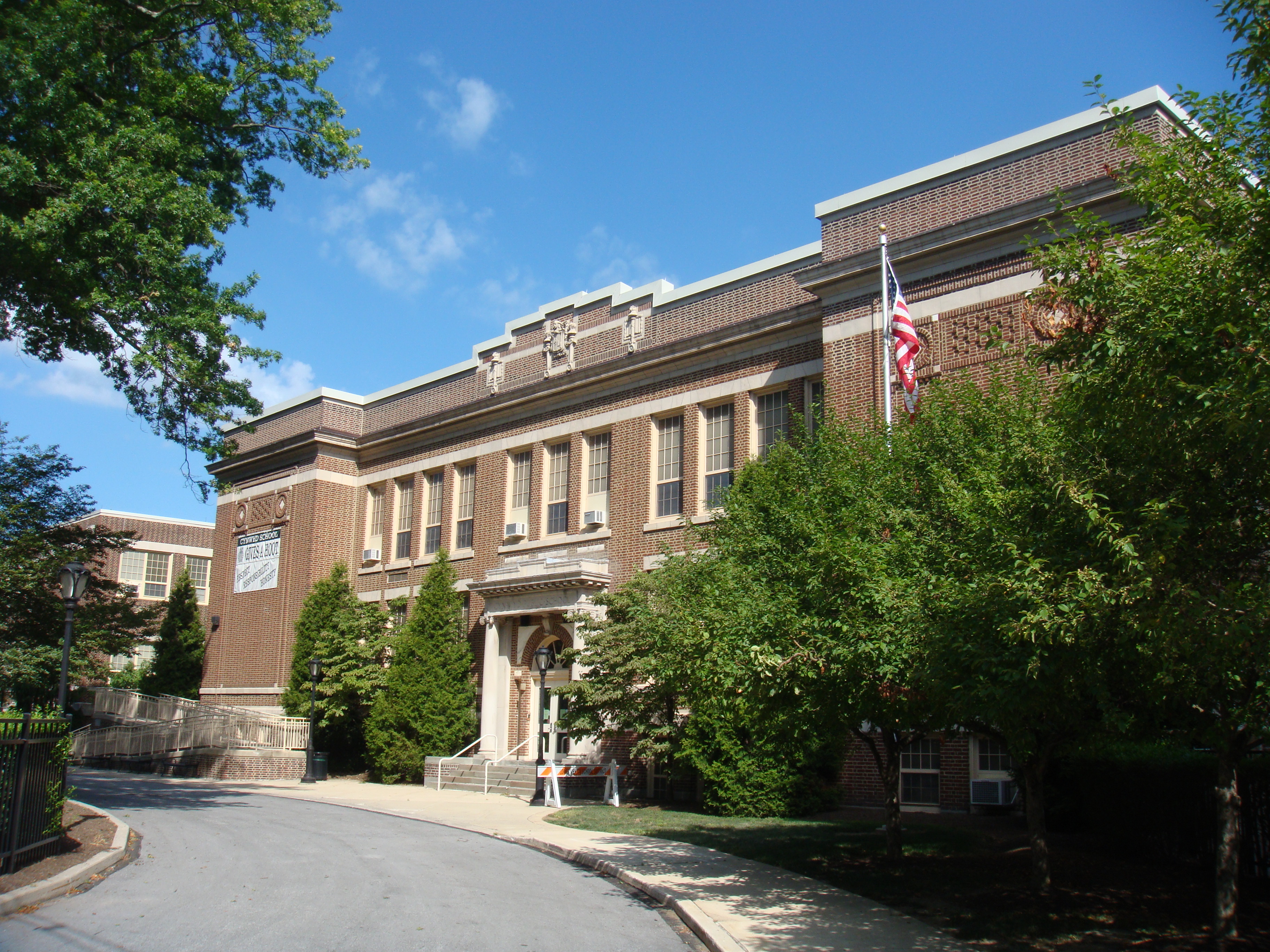 Multi-story brick school building with columns, flagpole, and mature trees under blue sky.
