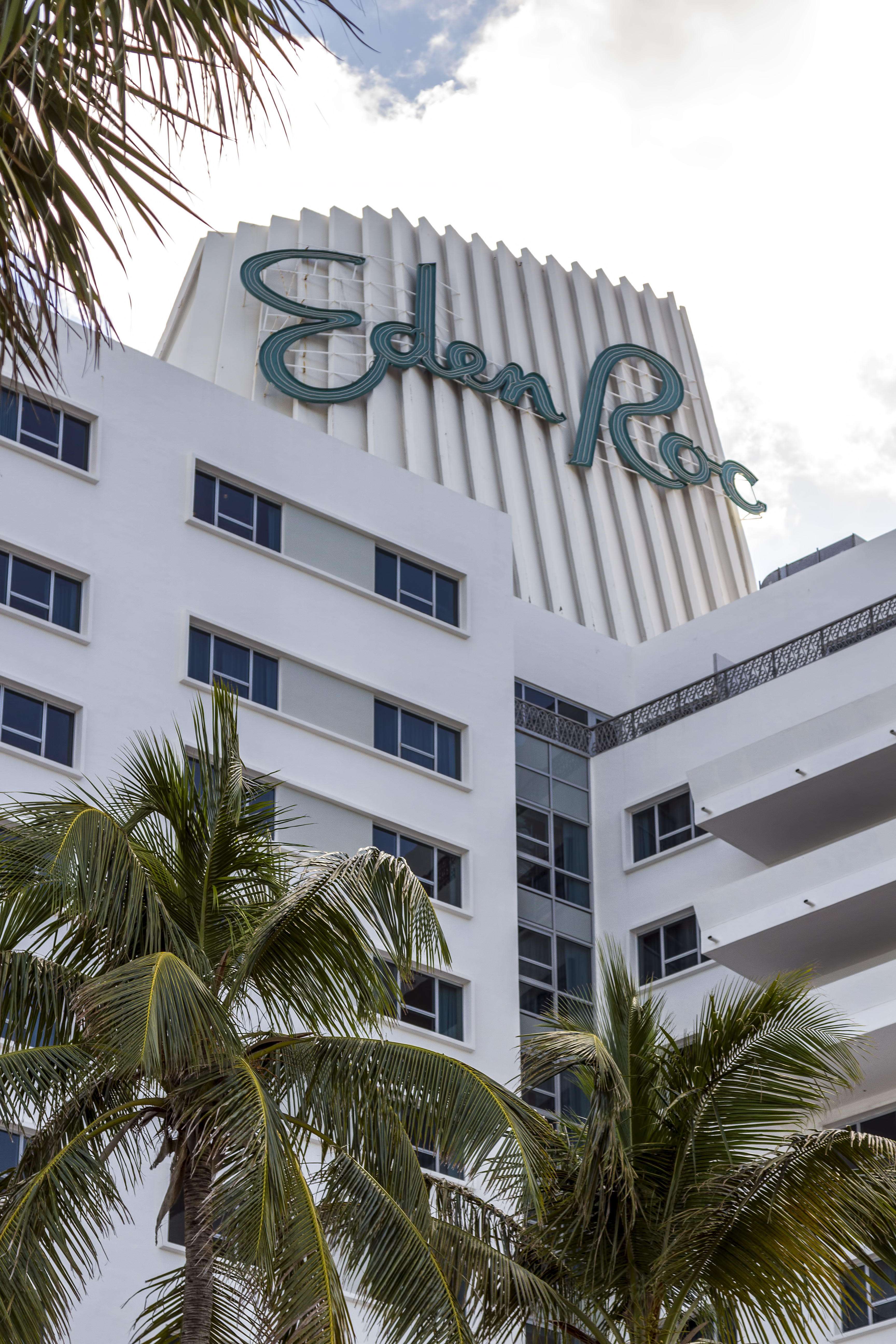 White Art Deco hotel building with teal cursive sign and palm trees in the foreground.