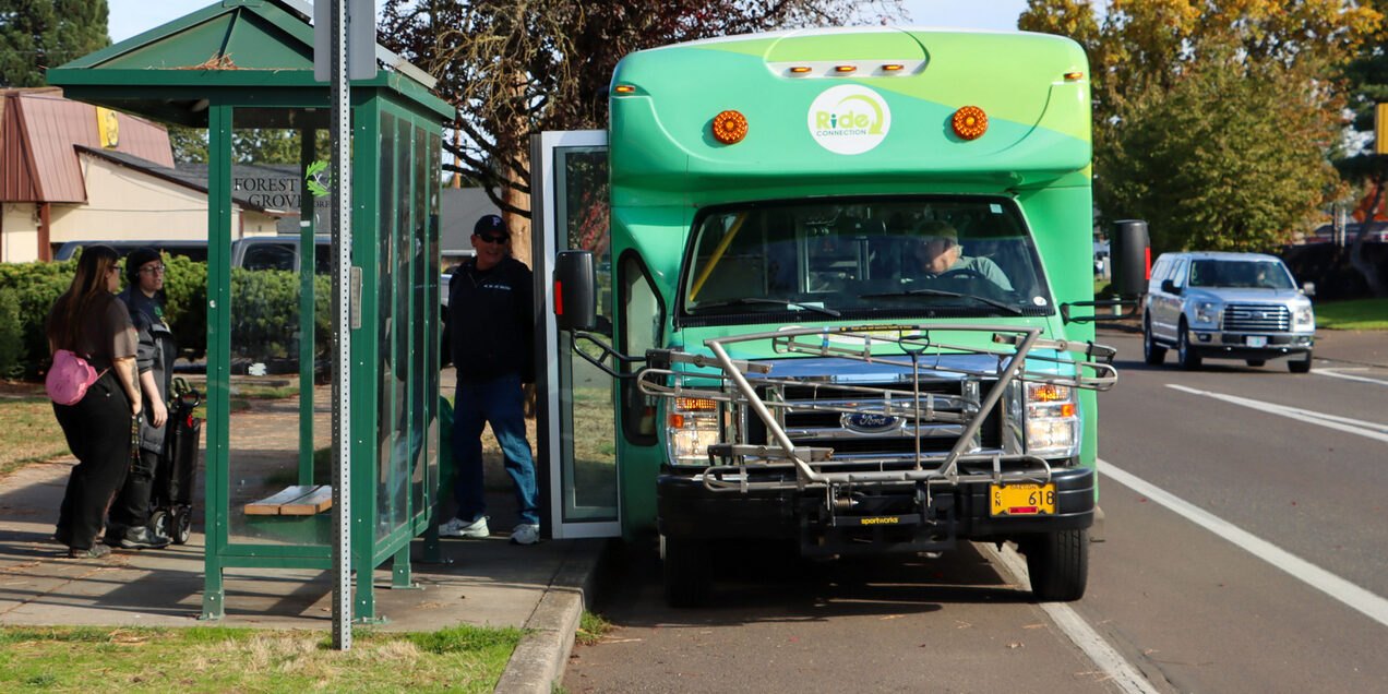 Passengers boarding a small green transit shuttle bus at a covered bus shelter.