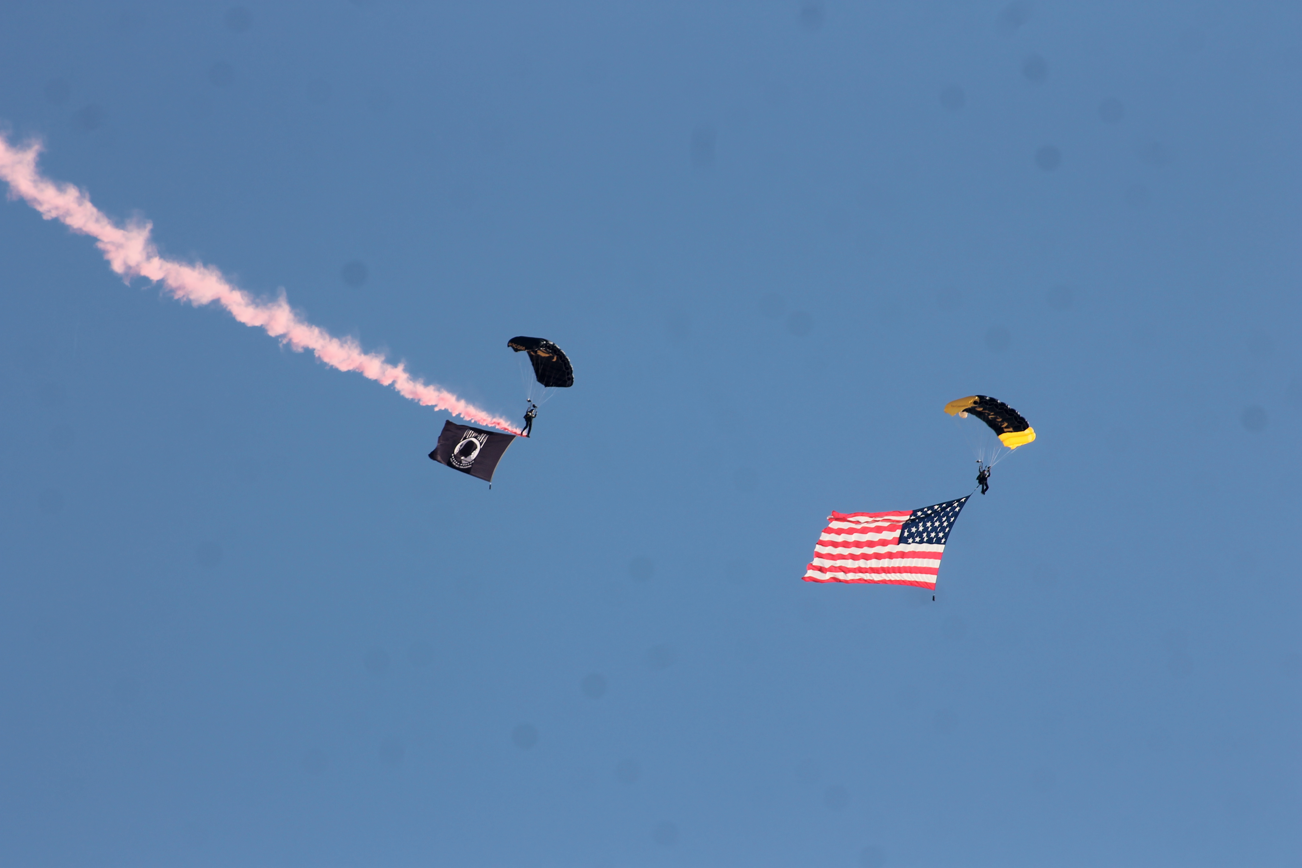 Two paratroopers under dark parachutes carry American and POW/MIA flags through a blue sky.