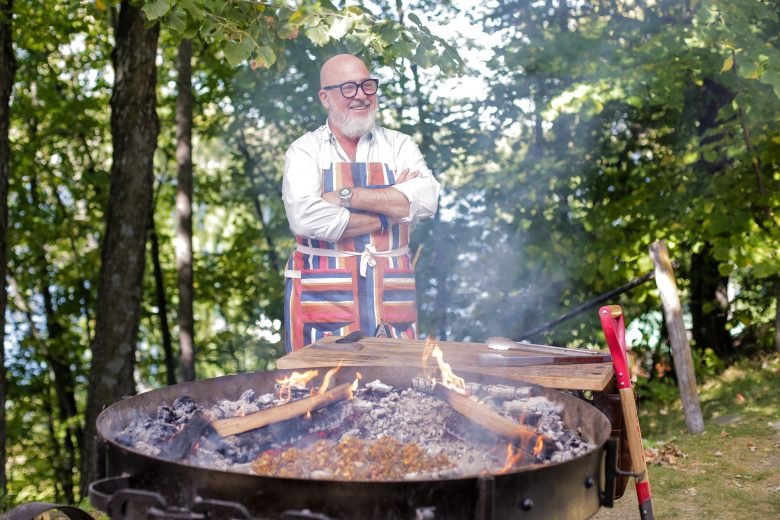 Man with white beard and glasses in a striped apron stands behind a large outdoor fire pit.
