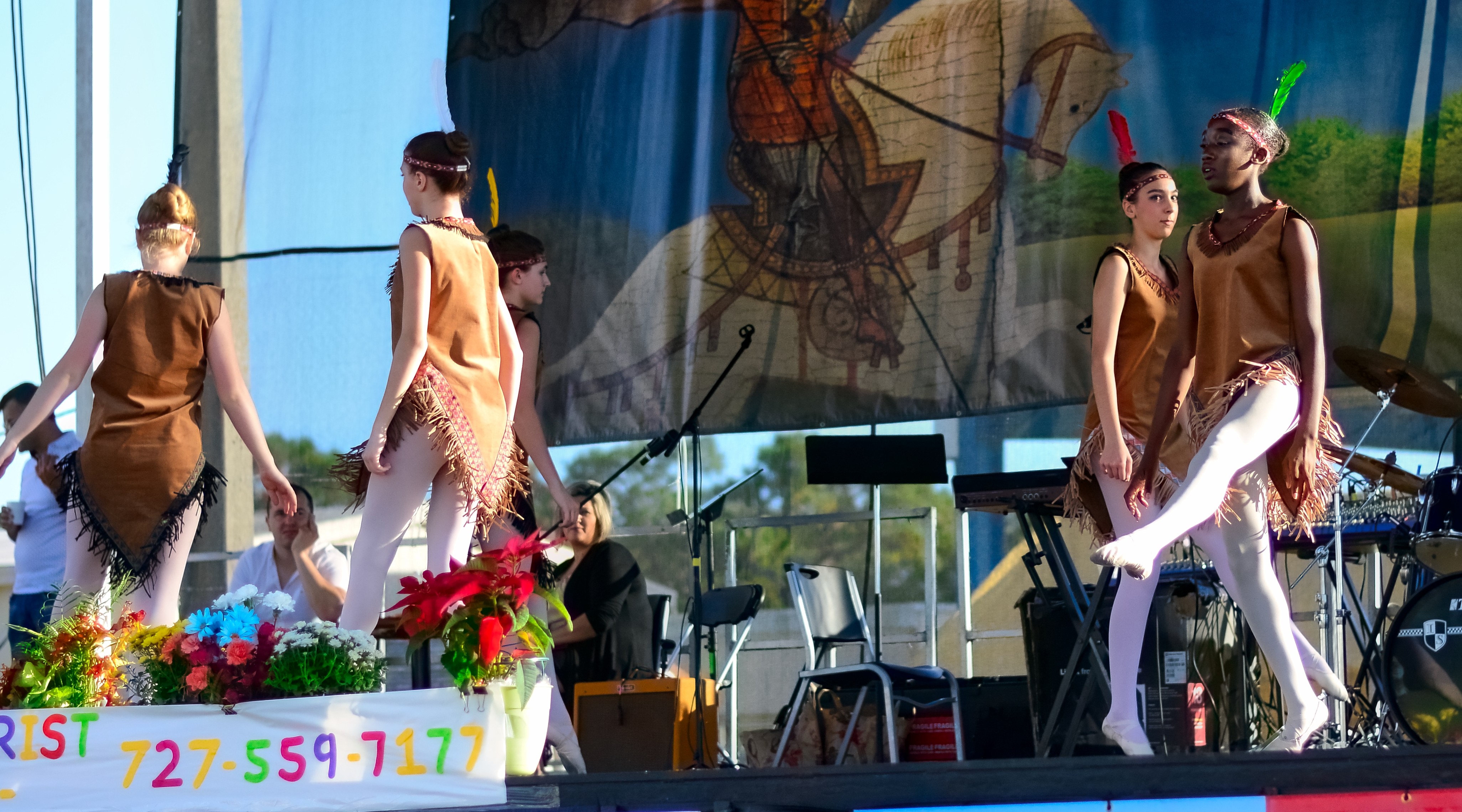 Ballet dancers in fringed costumes performing on an outdoor stage with a painted backdrop.