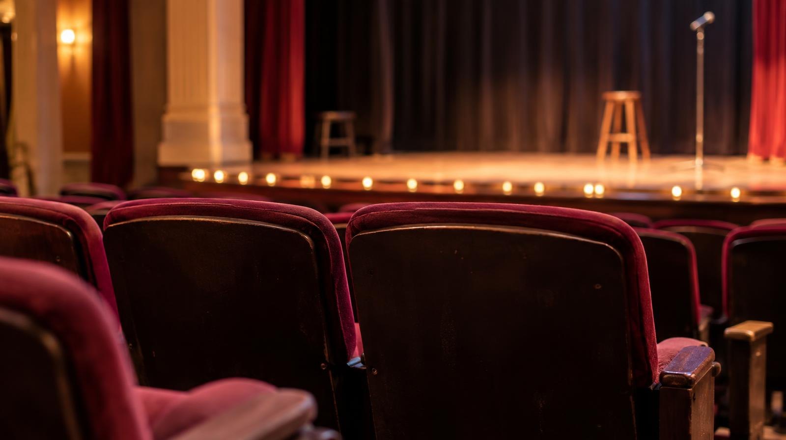 Empty red theater seats face a wooden stage lit by warm footlights.