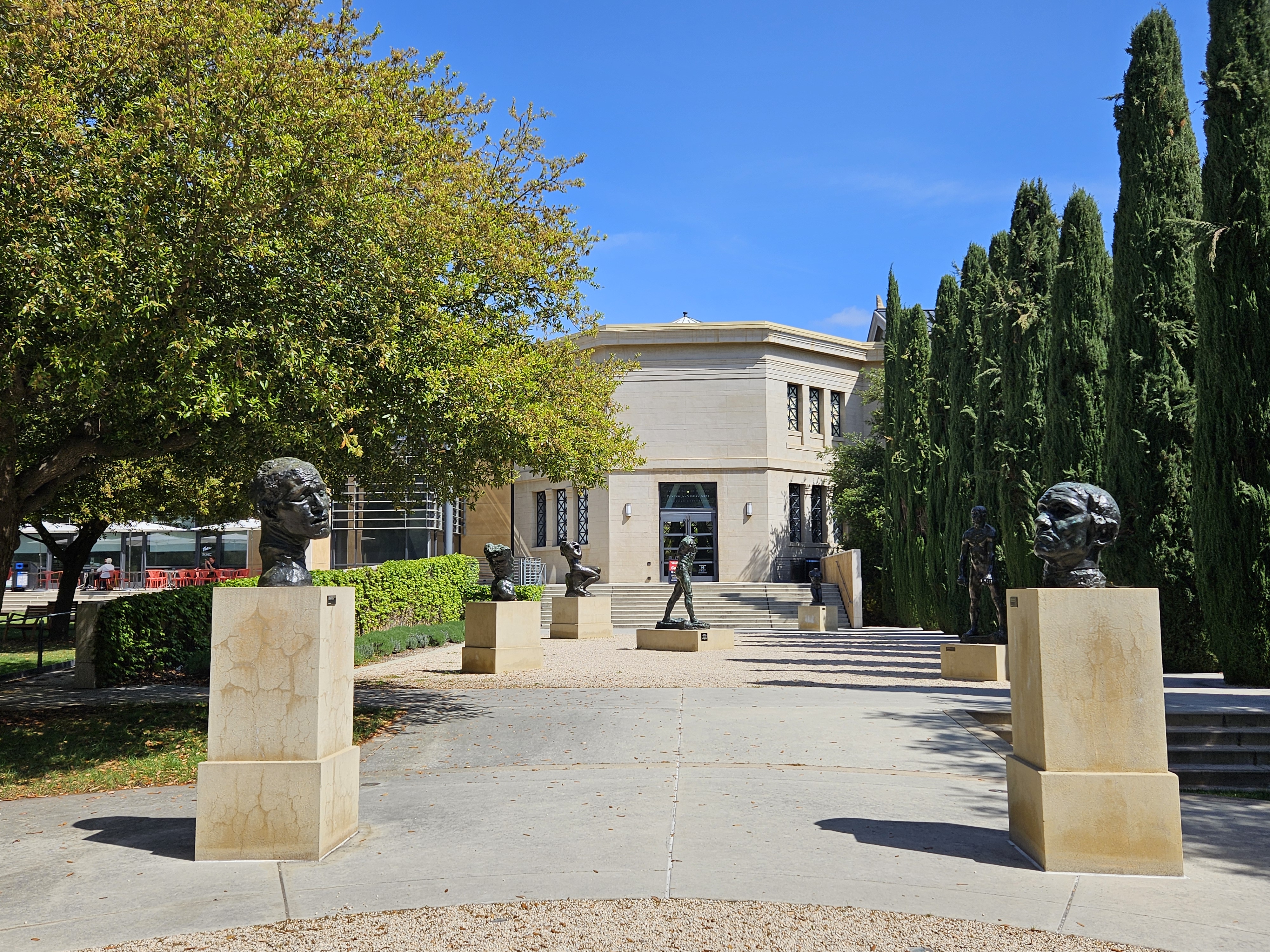 Bronze sculptures on pedestals line a cypress-flanked walkway toward a neoclassical building.