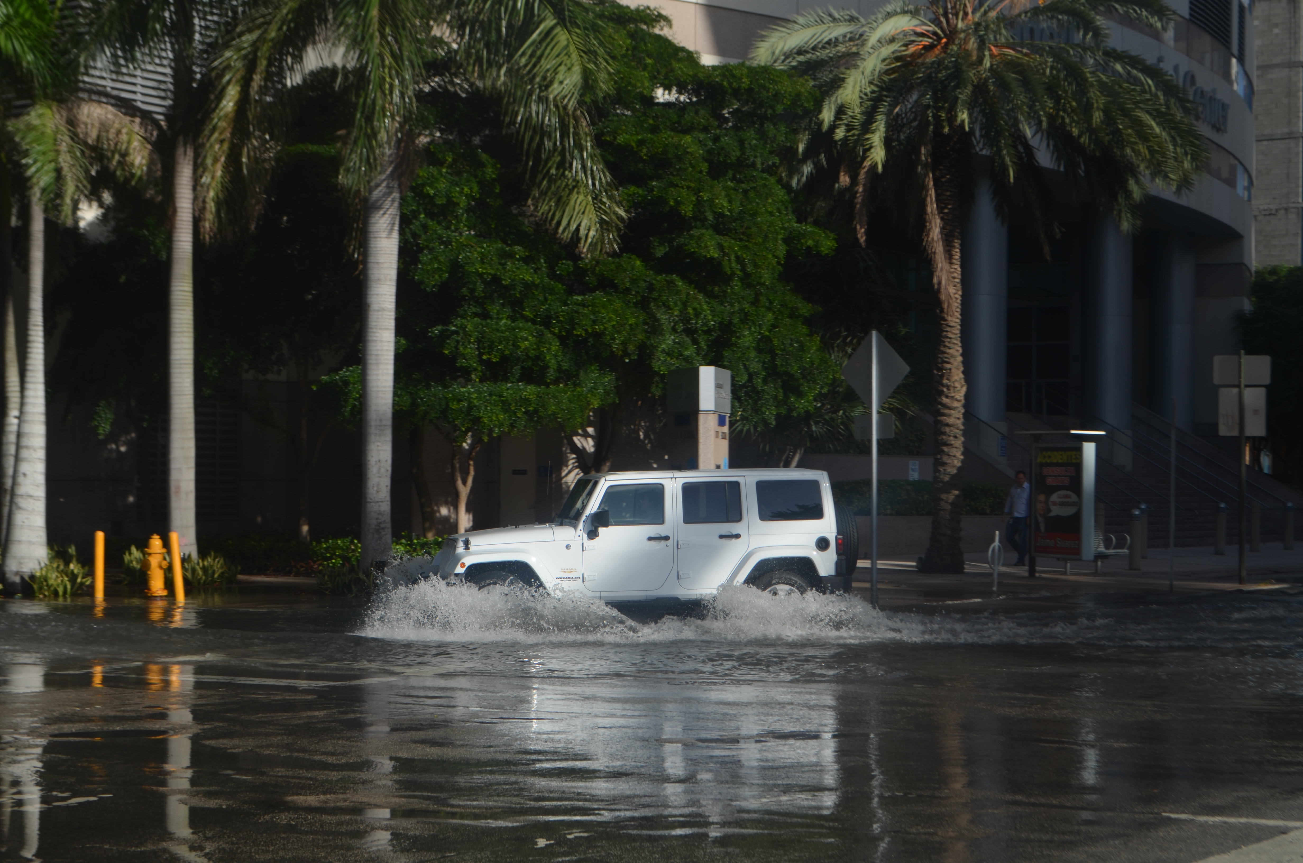 An SUV drives through deep floodwater on a city street, water splashing around its tires.