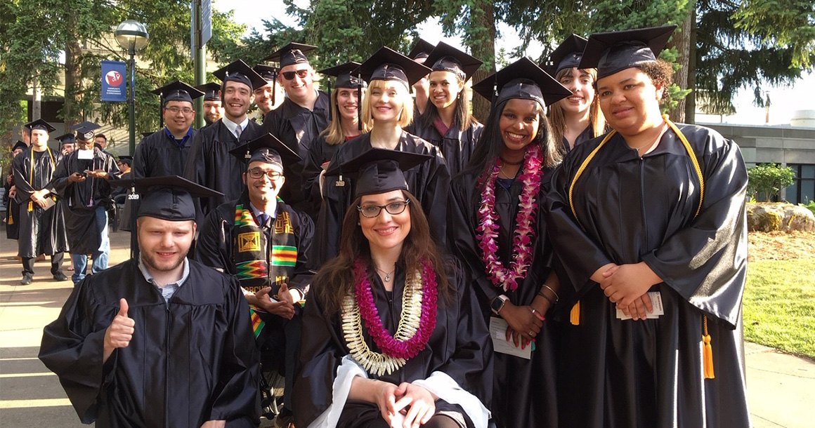 A large group of students in black graduation caps and gowns pose outdoors.