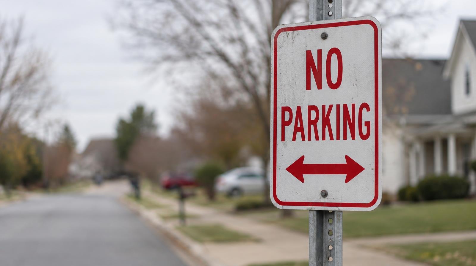 A red and white no-parking sign on a metal pole beside a quiet residential street.