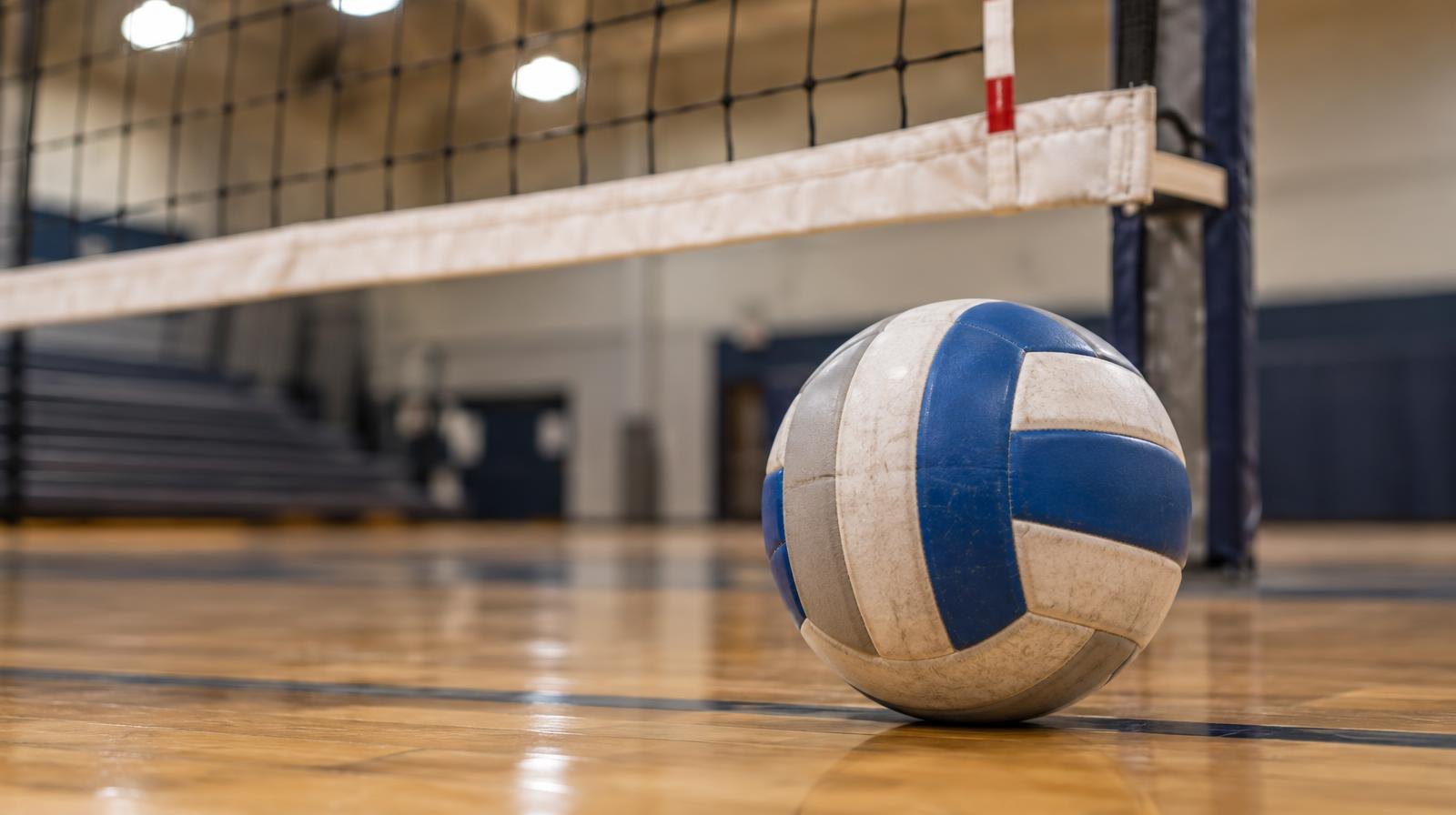 A volleyball resting on a hardwood floor beside a volleyball net.