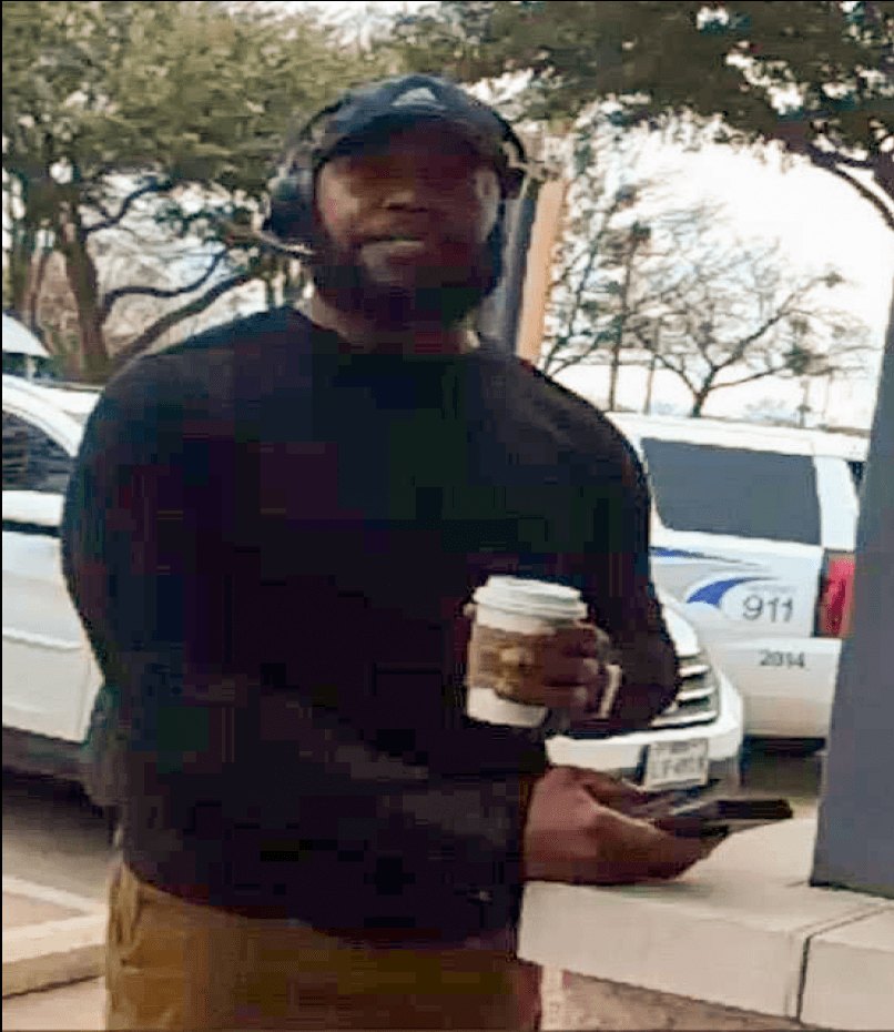 Man in plaid shirt and baseball cap holding a coffee cup near a police vehicle marked 911.
