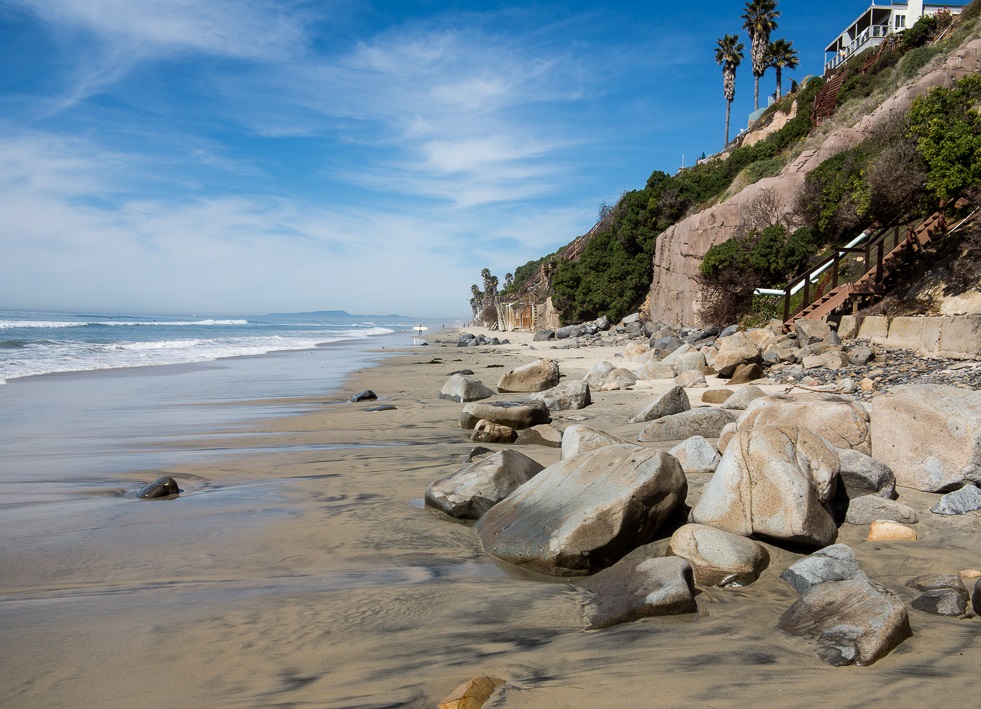 Rocky beach with sandstone bluffs, palm trees, and a wooden staircase descending the cliff.
