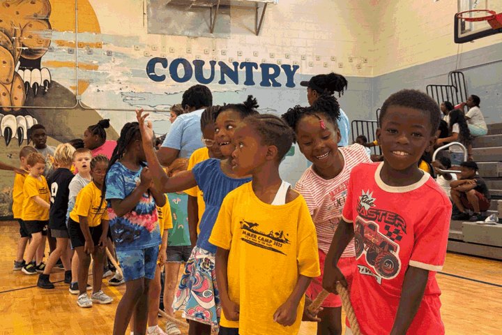 Children stand together inside a gymnasium with a painted mural on the wall.