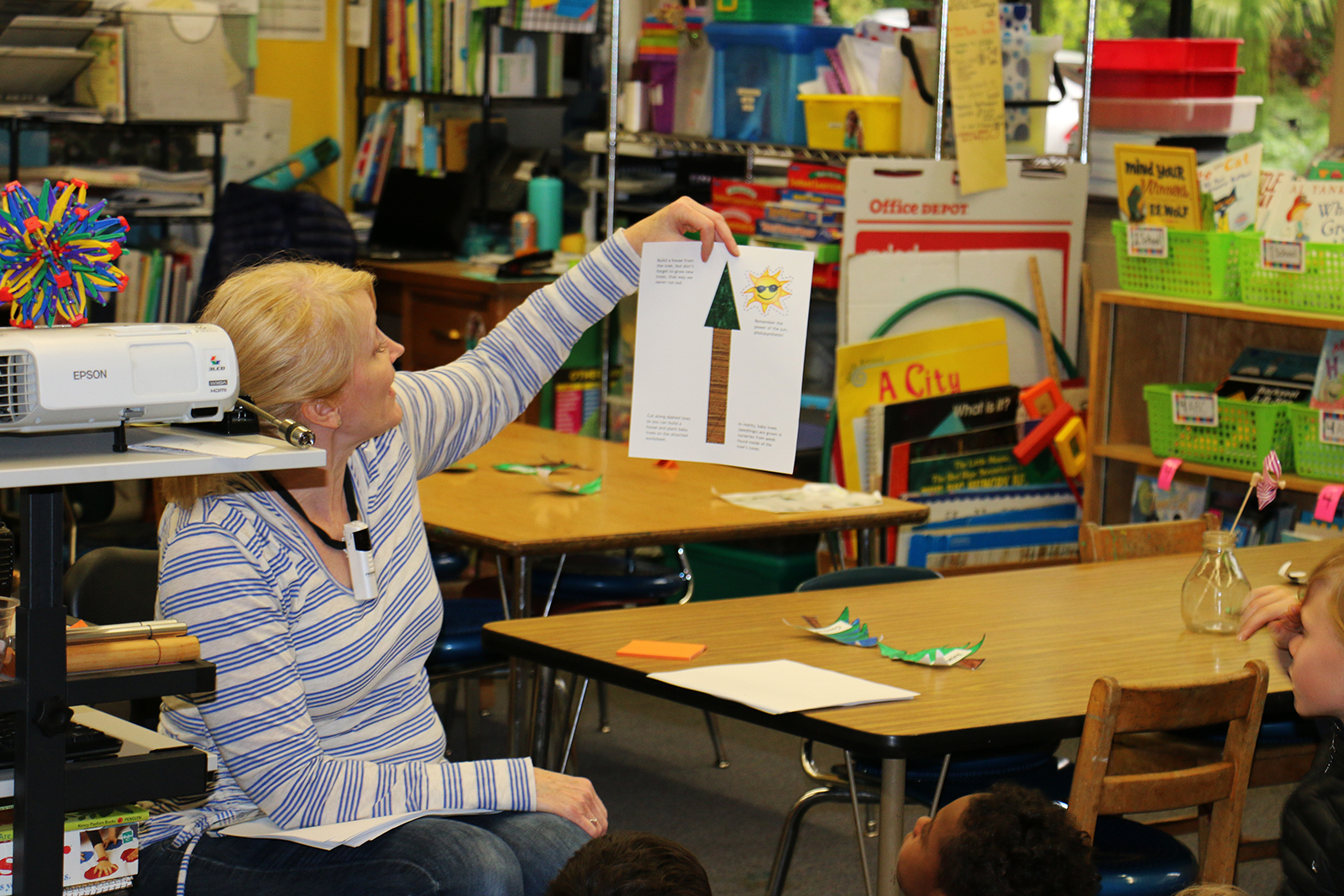 A teacher holds up a printed page while students sit in an elementary school classroom.