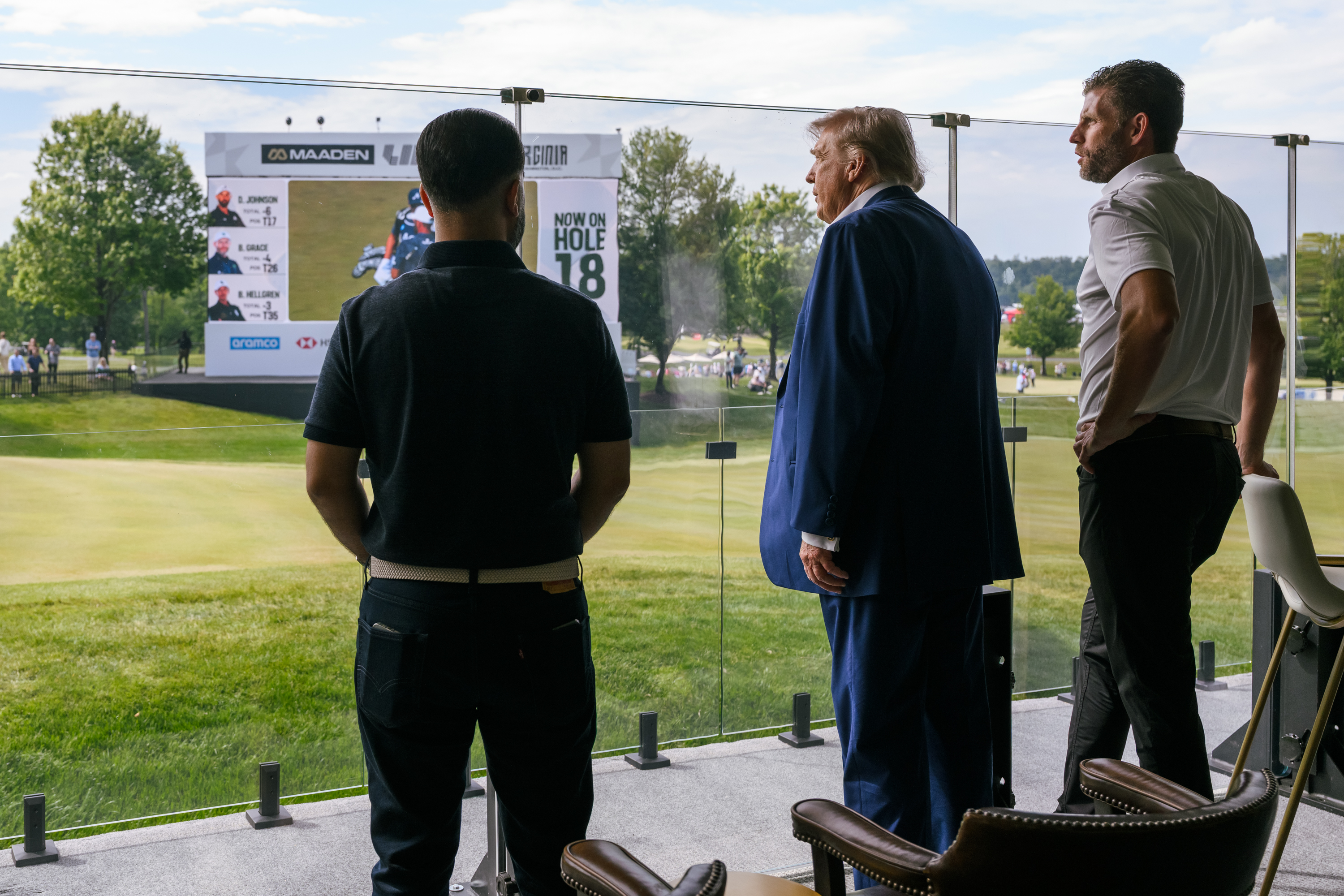 Three men stand on a viewing platform overlooking a golf course with a LIV Golf scoreboard.