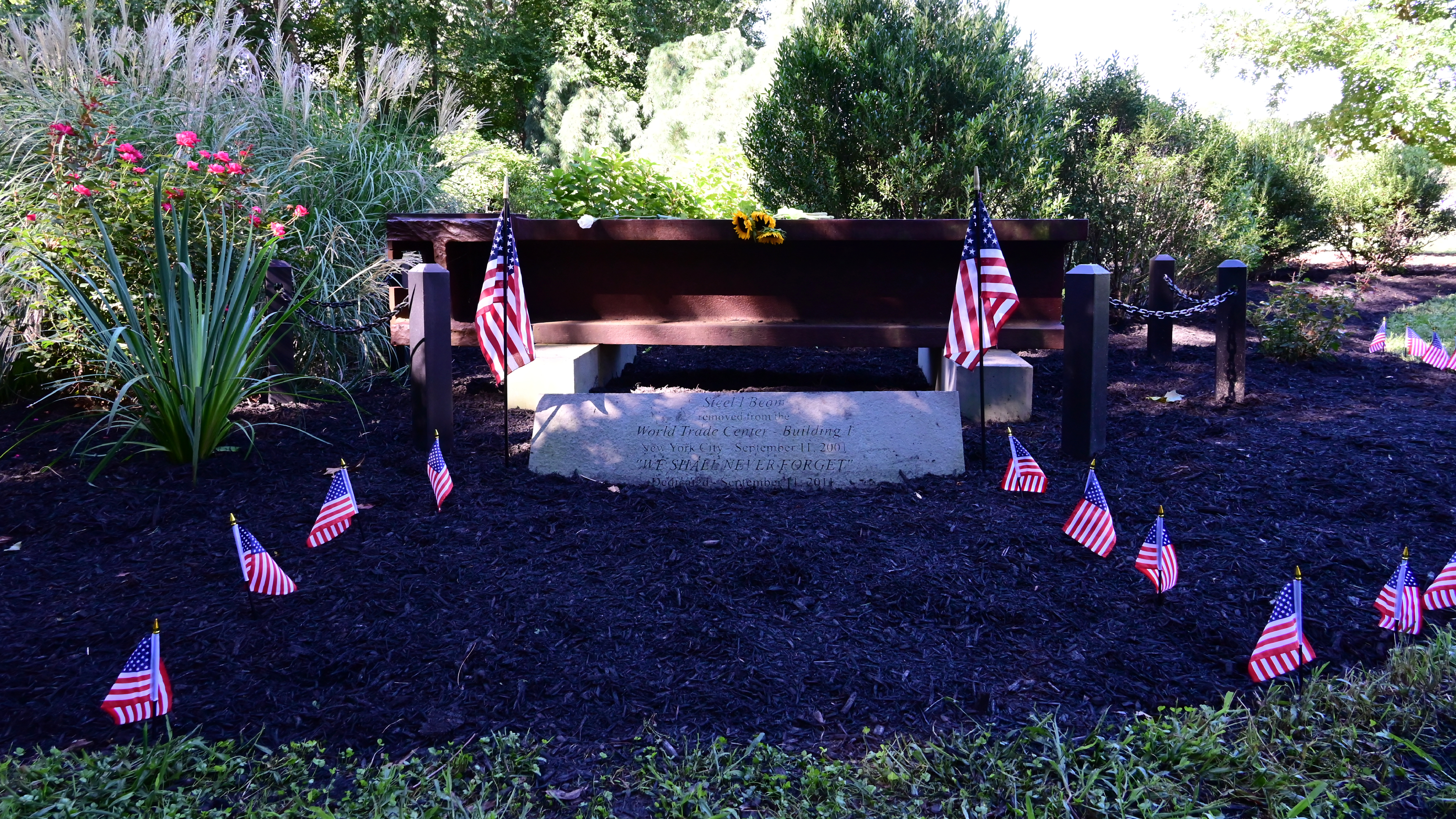 Small American flags planted around a stone 9/11 memorial marker in a garden setting.