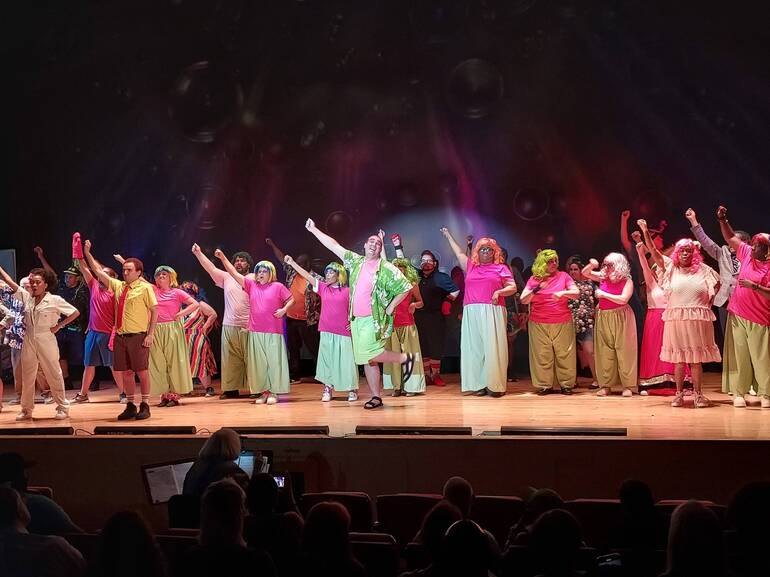 Large ensemble cast in bright costumes with arms raised on a colorfully lit theater stage.