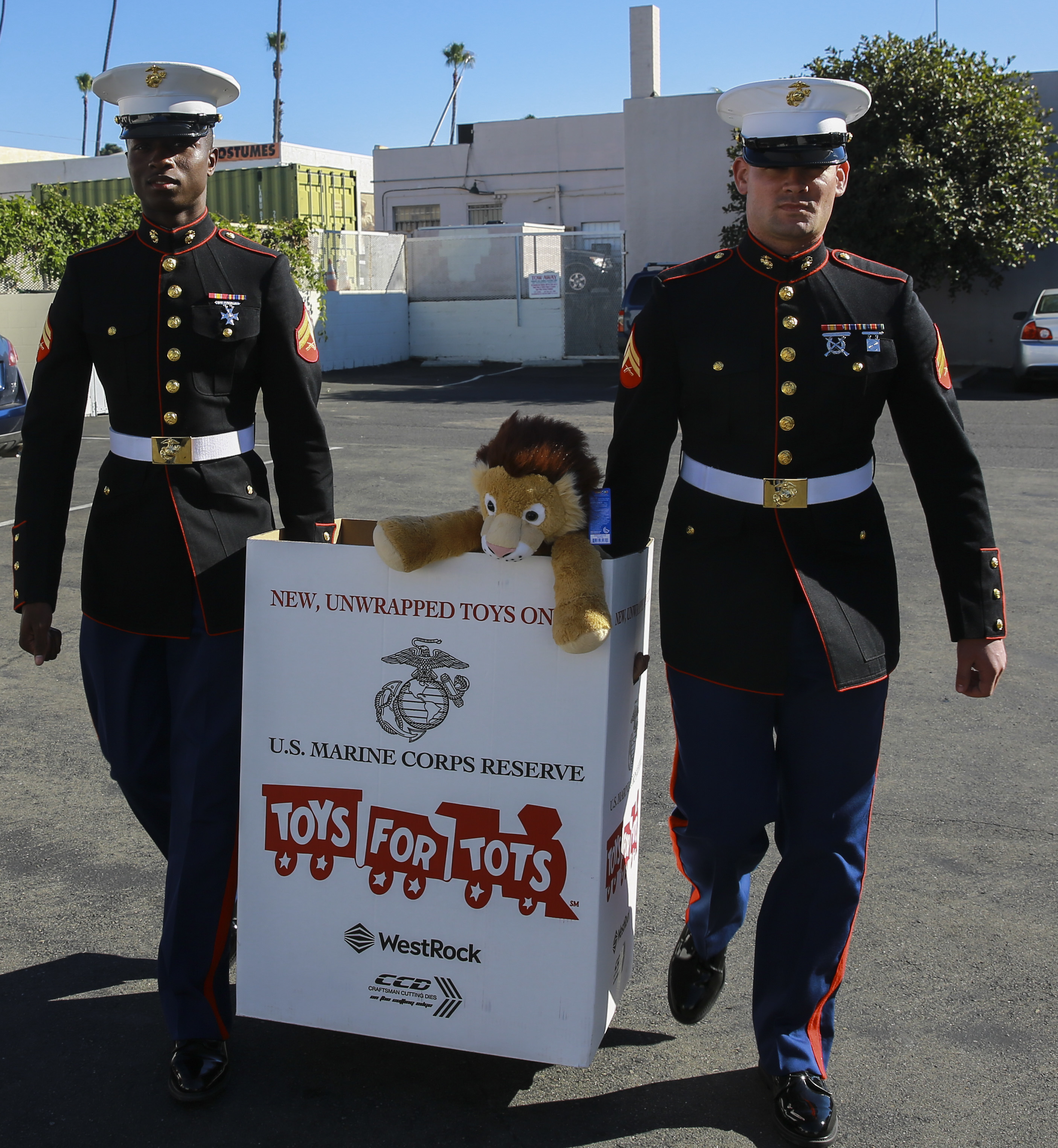 Two Marines in dress uniforms carry a branded Toys for Tots donation box.