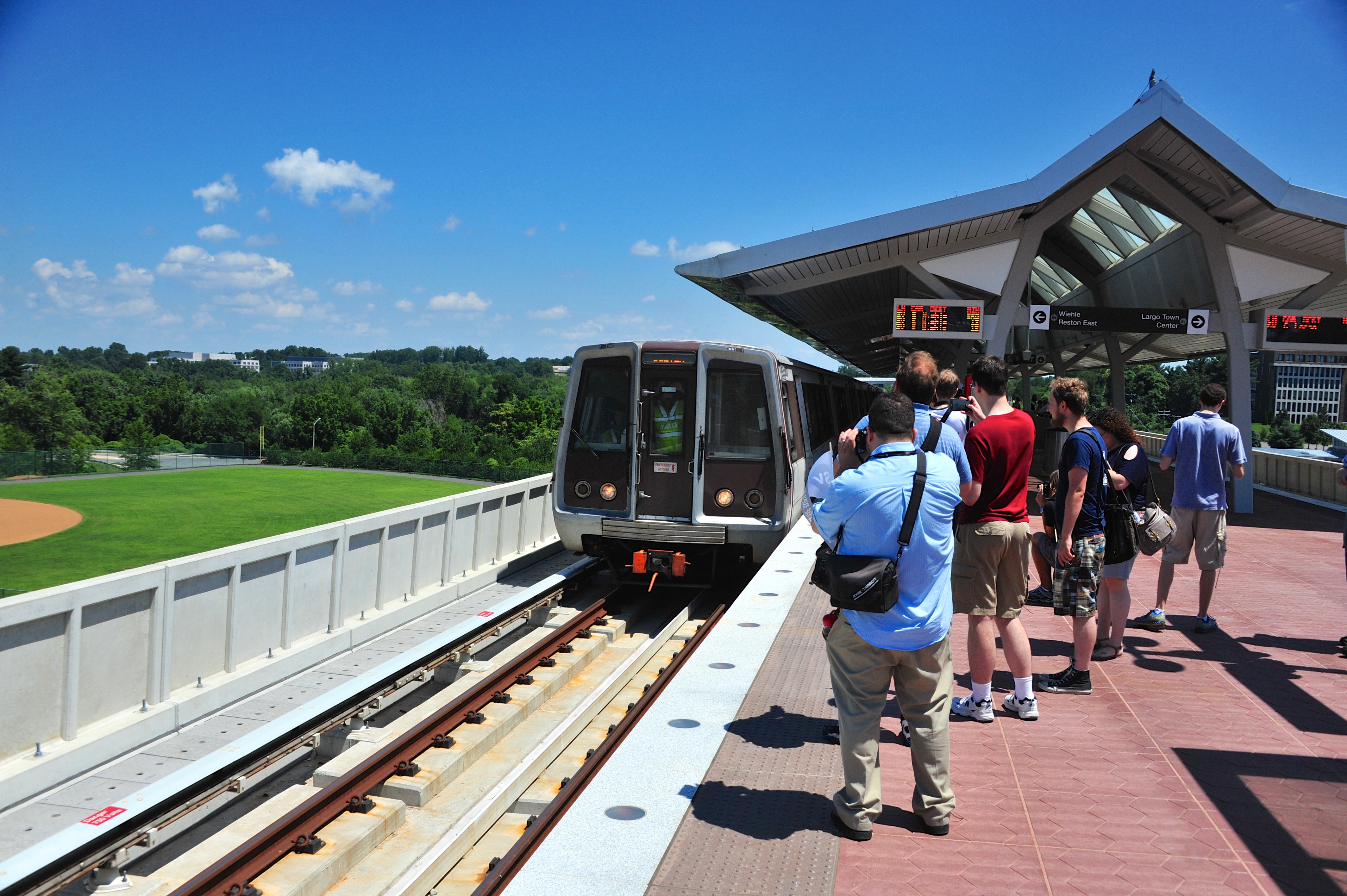 A Metro train arriving at an elevated station platform with passengers waiting under a peaked roof structure.