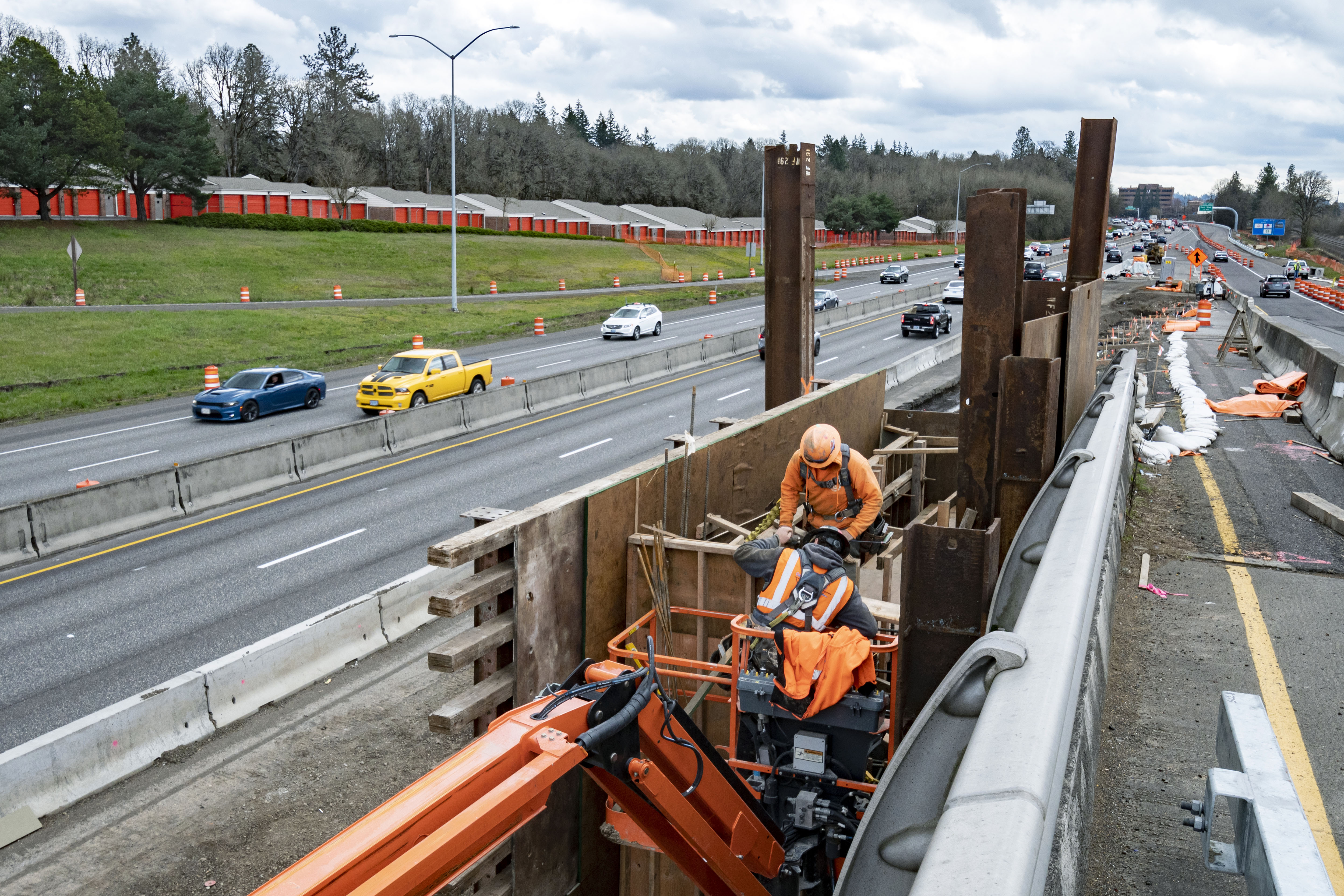 Construction worker in safety vest on a boom lift next to a highway overpass with traffic and orange cones below.