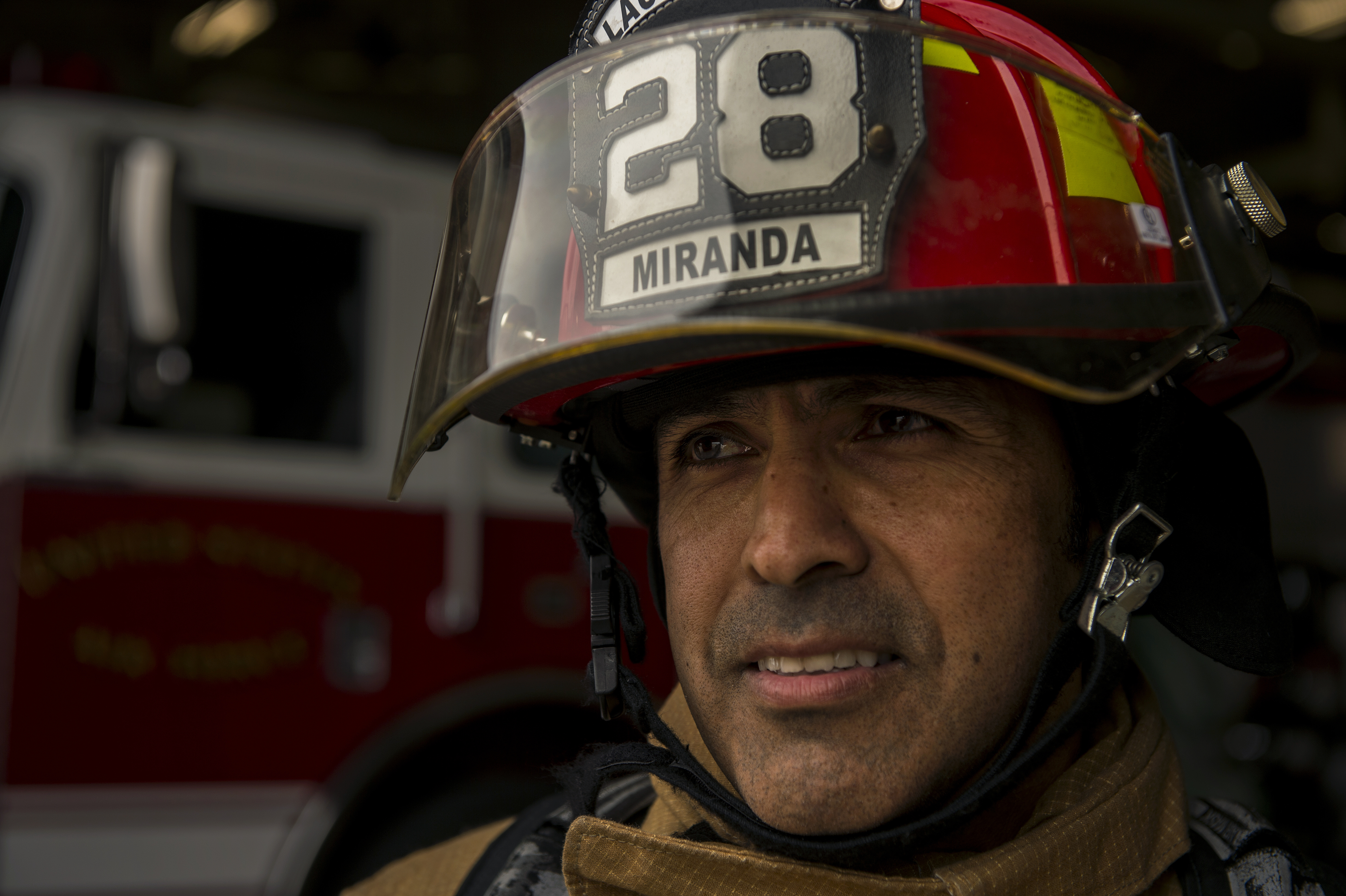 Firefighter in full turnout gear and red helmet stands in front of a fire truck.