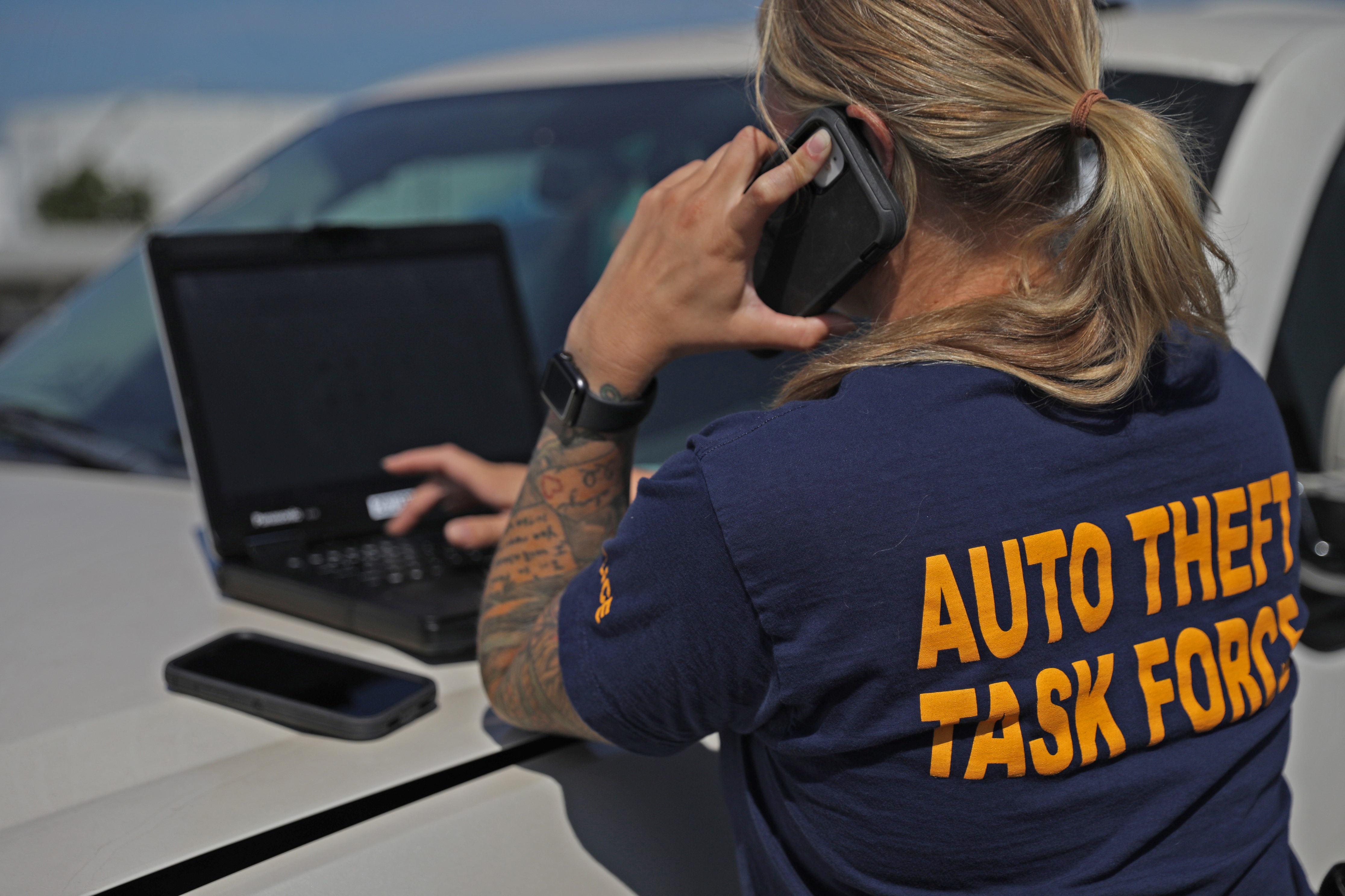 Person in Auto Theft Task Force shirt talks on phone while using a laptop on a car hood.