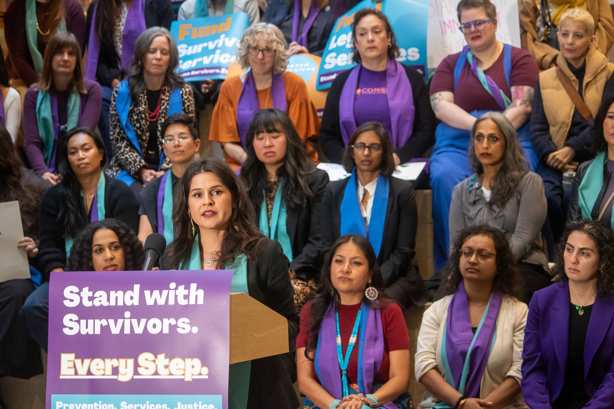 A speaker at a podium with a 'Stand with Survivors. Every Step.' sign, crowd behind her in orange, purple, and teal.