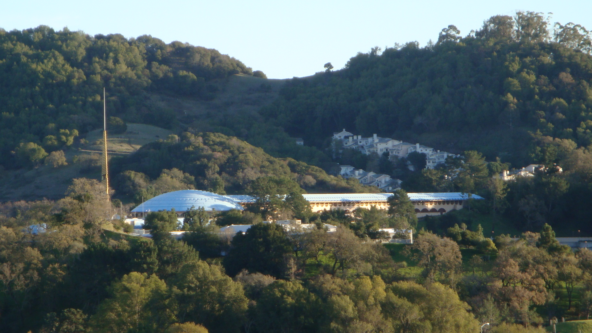 Curved civic building with a blue dome roof nestled among forested hills under soft light.