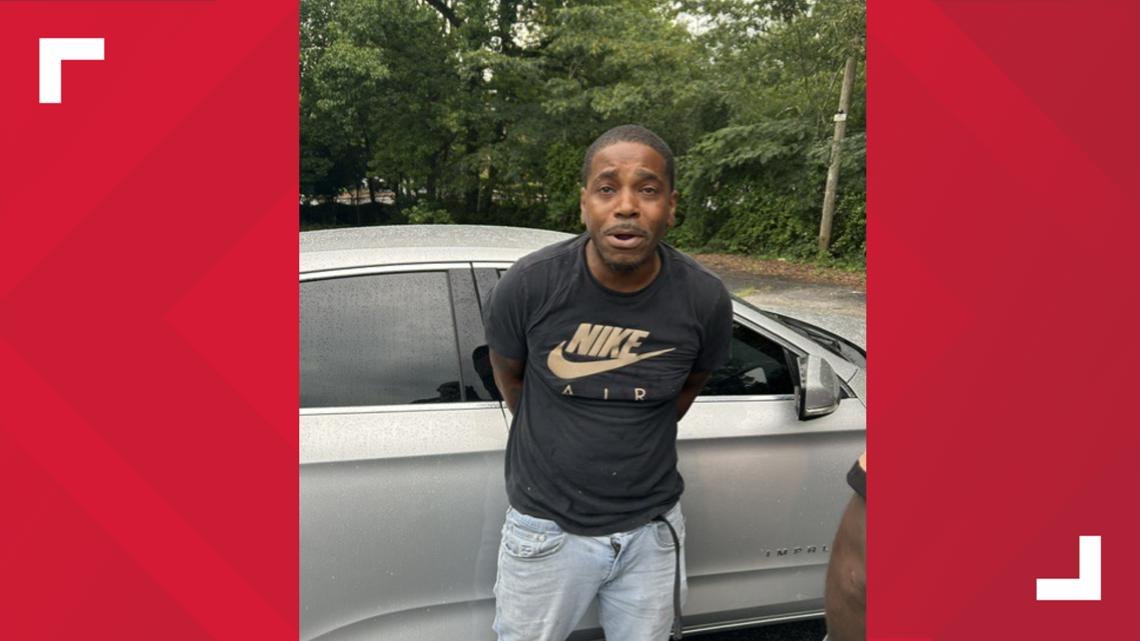 Man in a black t-shirt and jeans standing in front of a silver car outdoors.