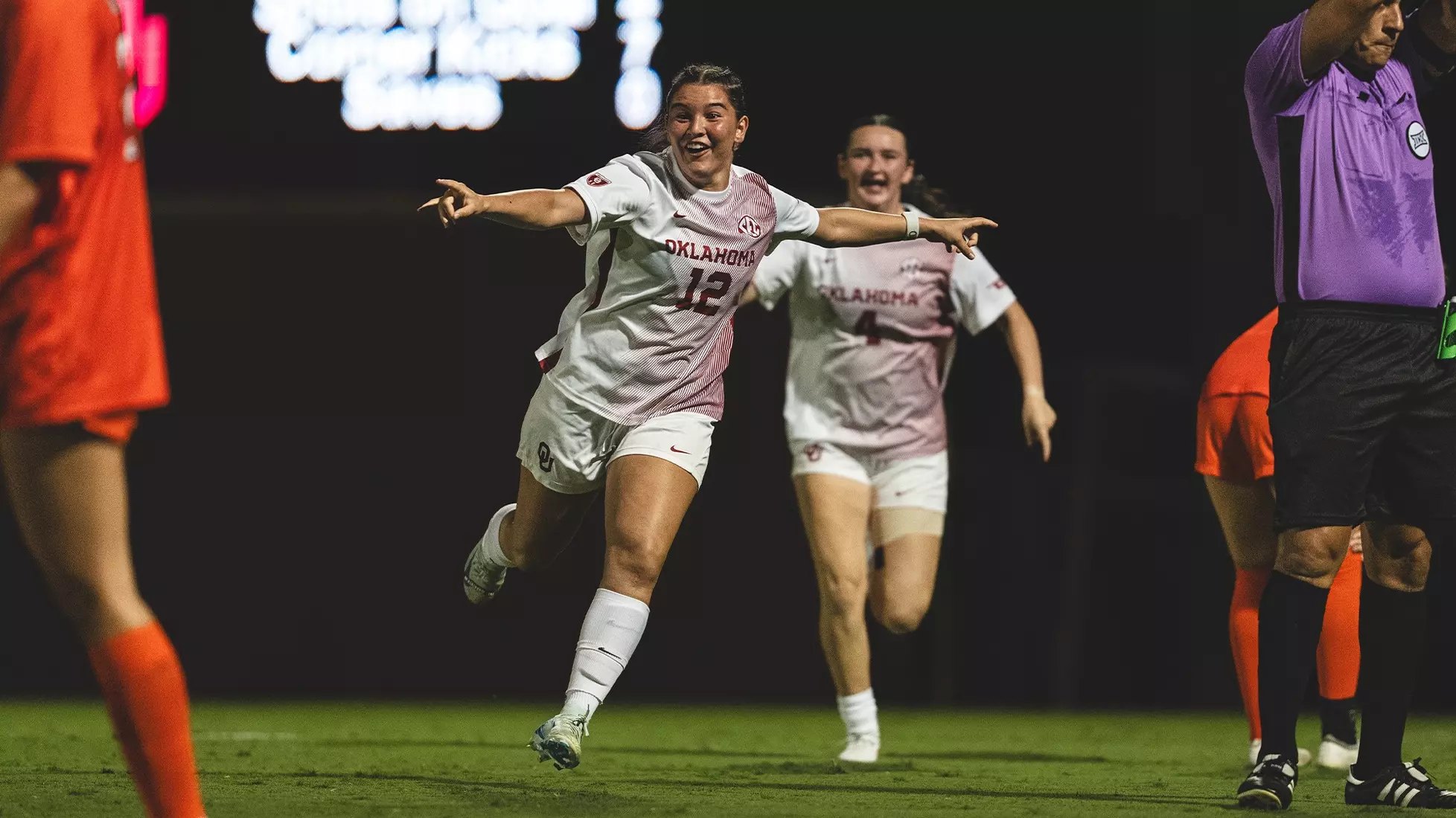 A soccer player in a white uniform celebrates with arms outstretched on a night field.