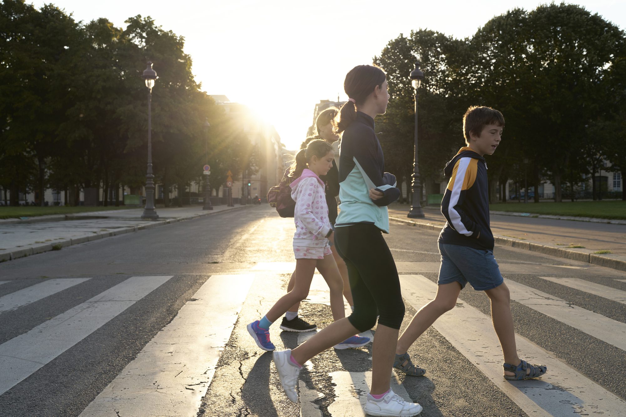 Group of people made up of children and adults crossing the street on the crosswalk, in the background there are trees and public lighting.