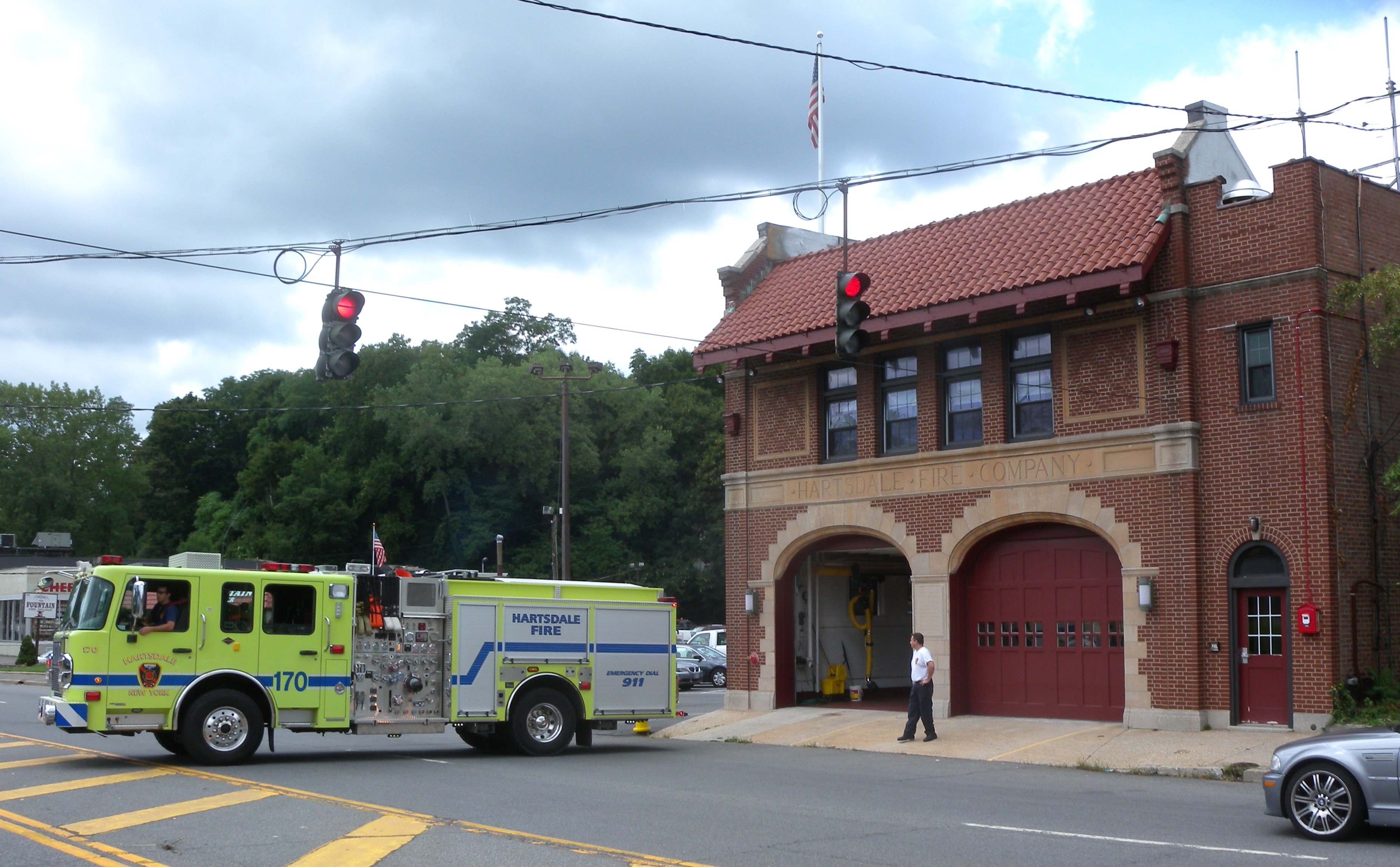 Yellow fire engine backing into a red-brick firehouse at a street corner.