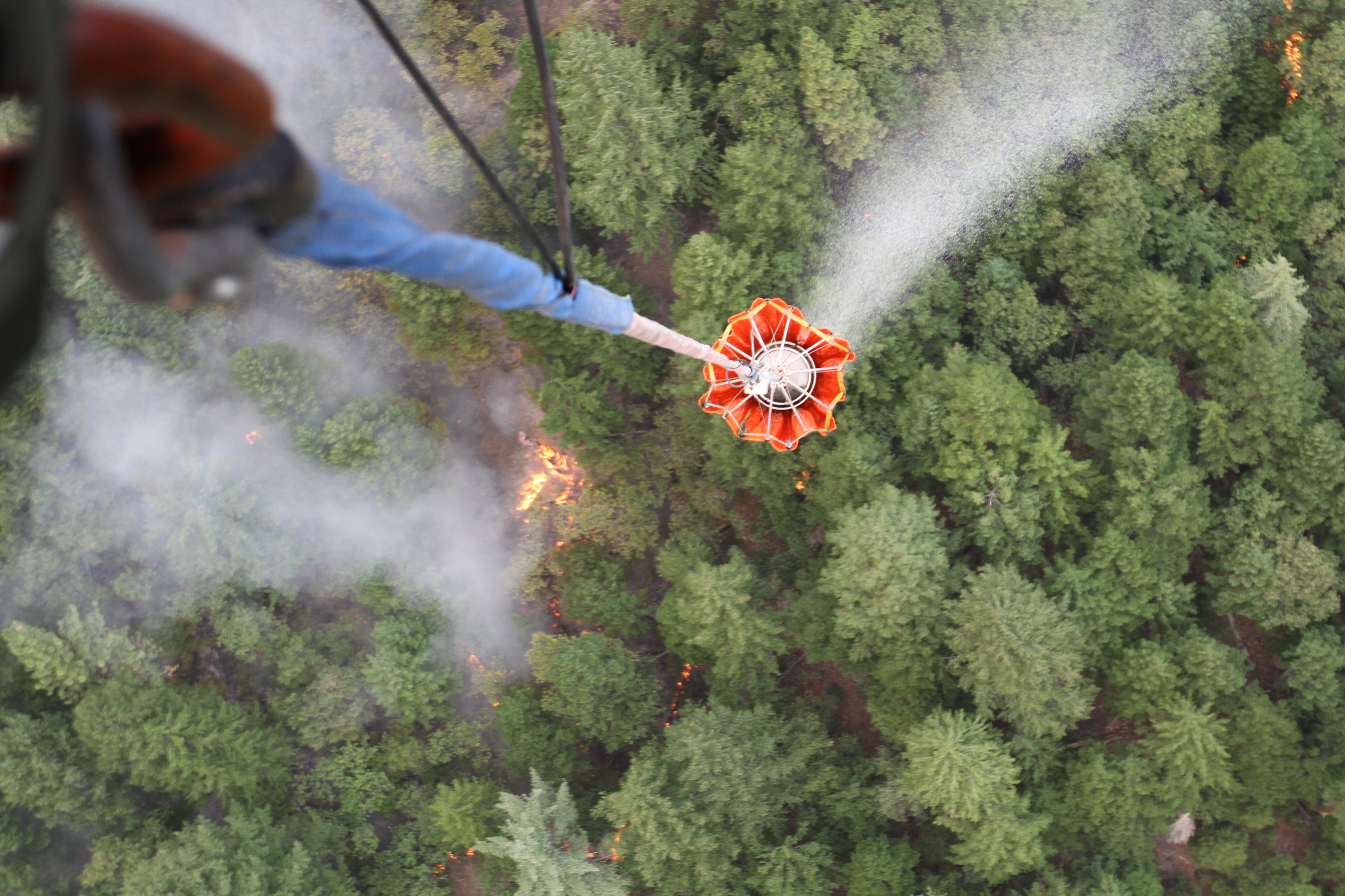 Aerial view of an orange water bucket dropping water over a forest with smoke and fire below.