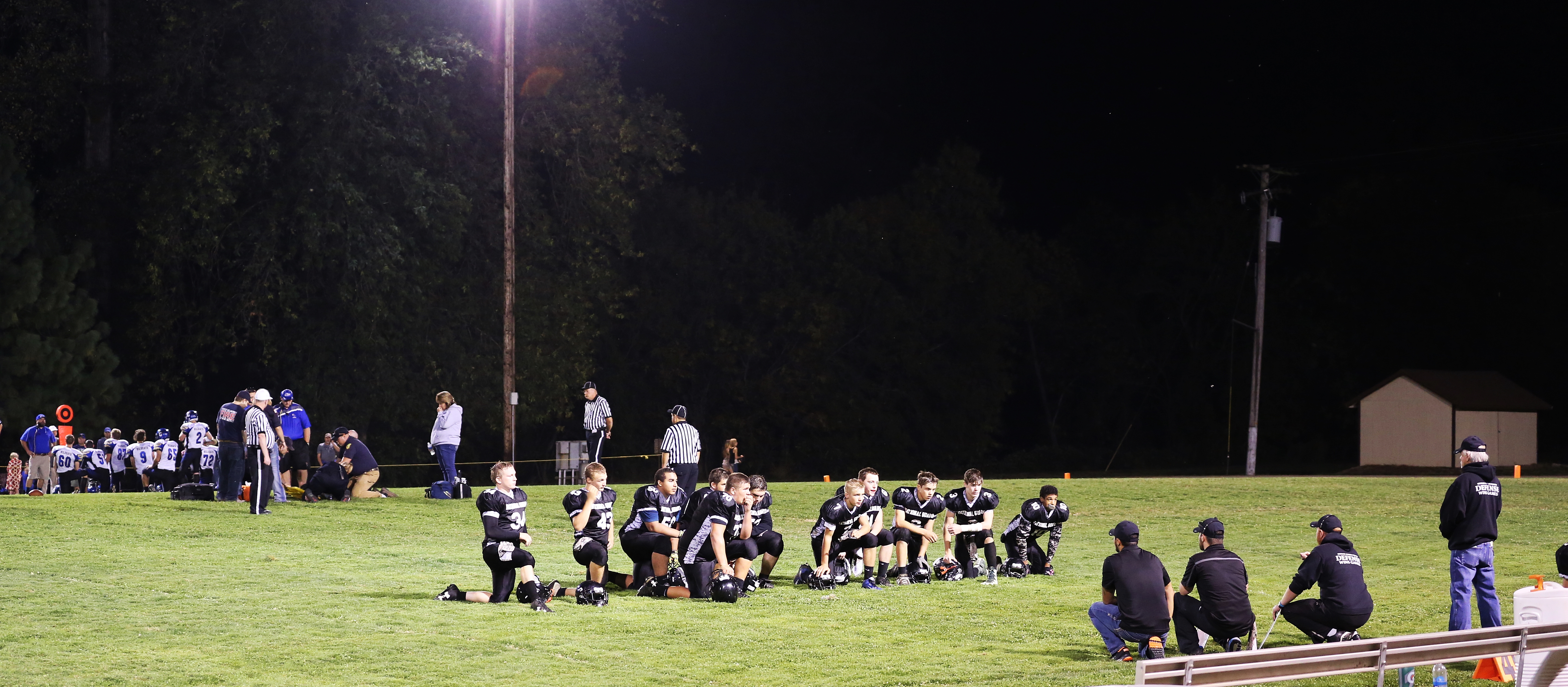 High school football players and coaches standing on a grass sideline under stadium lights at night.