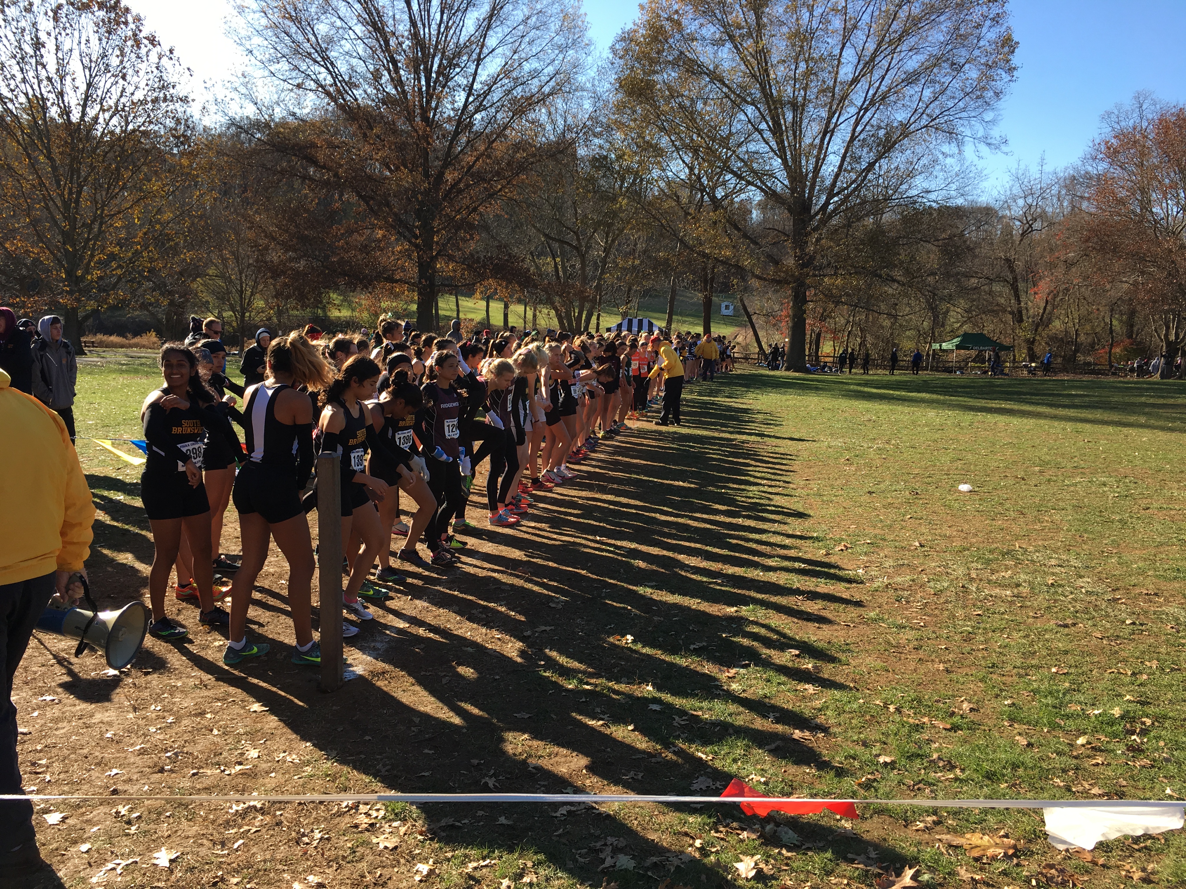Female runners lined up at the starting line of a high school cross country race on a grass field.