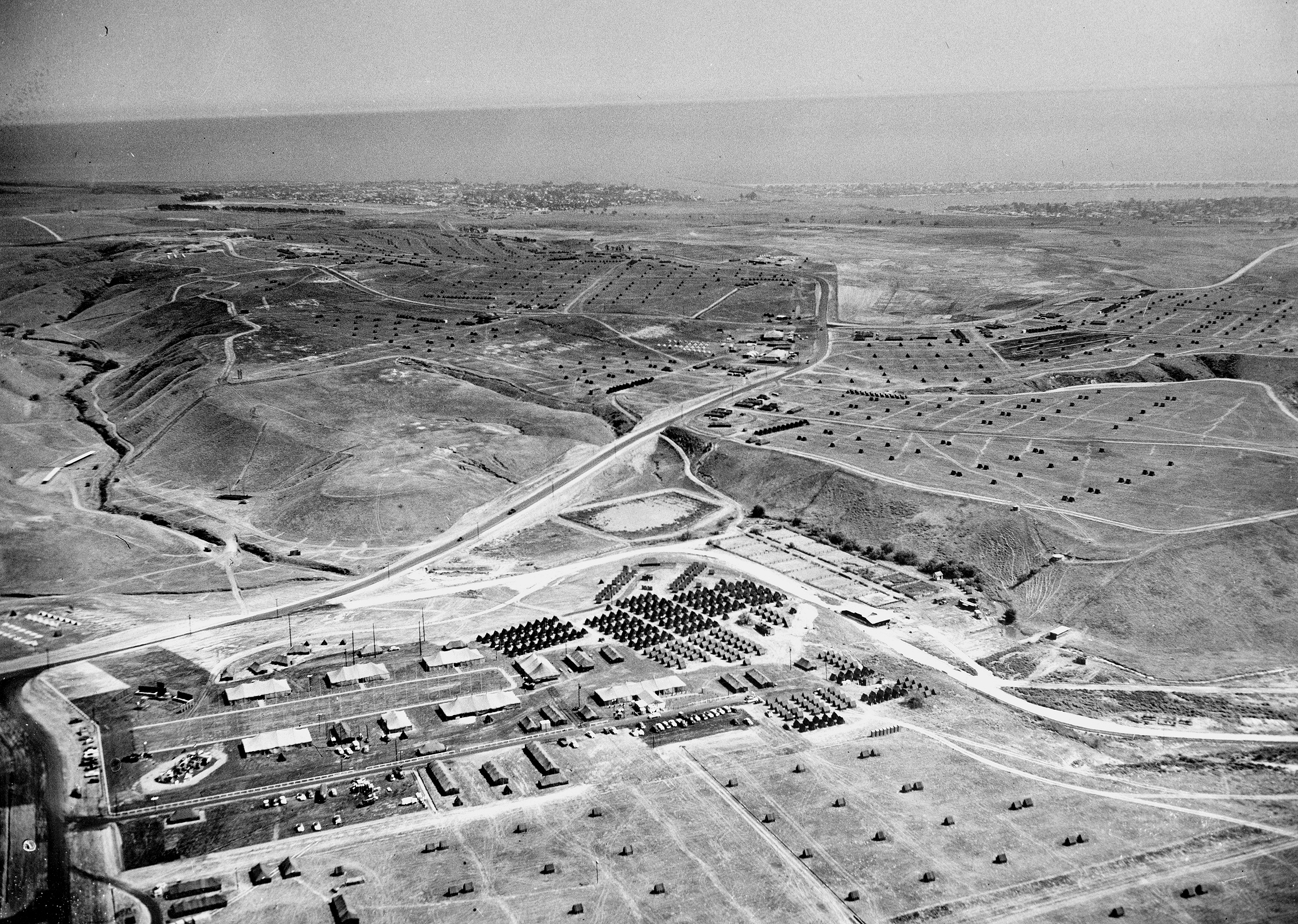 Aerial black-and-white photo of rows of tents at a large Boy Scout encampment on hilly terrain near the ocean.