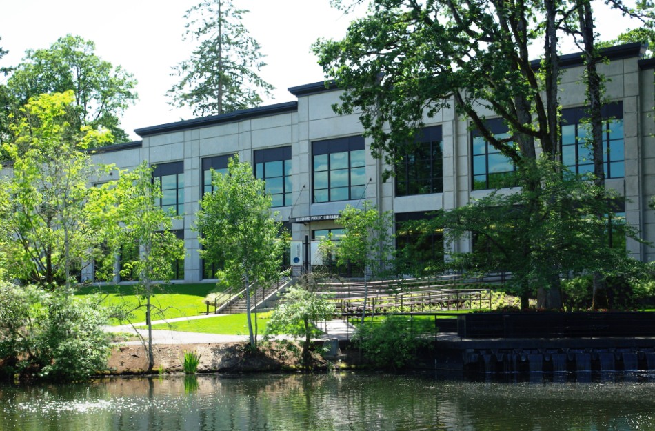 Modern two-story library building with large windows beside a pond and pedestrian bridge.