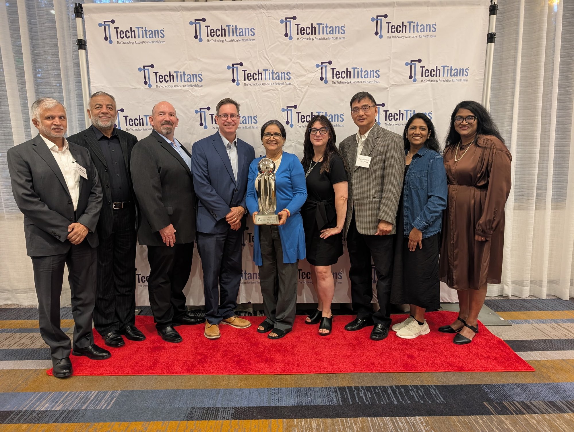 Nine people stand on a red carpet in front of a Tech Titans banner, one holding a silver trophy.