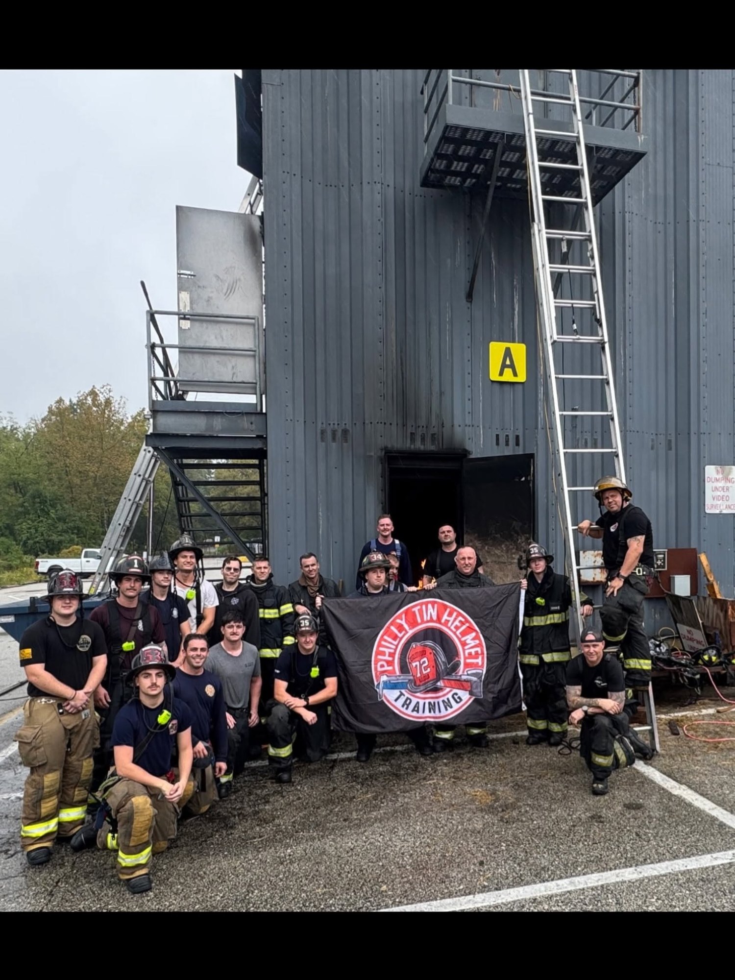 Firefighters in turnout gear and helmets pose in front of a metal training tower holding a banner.