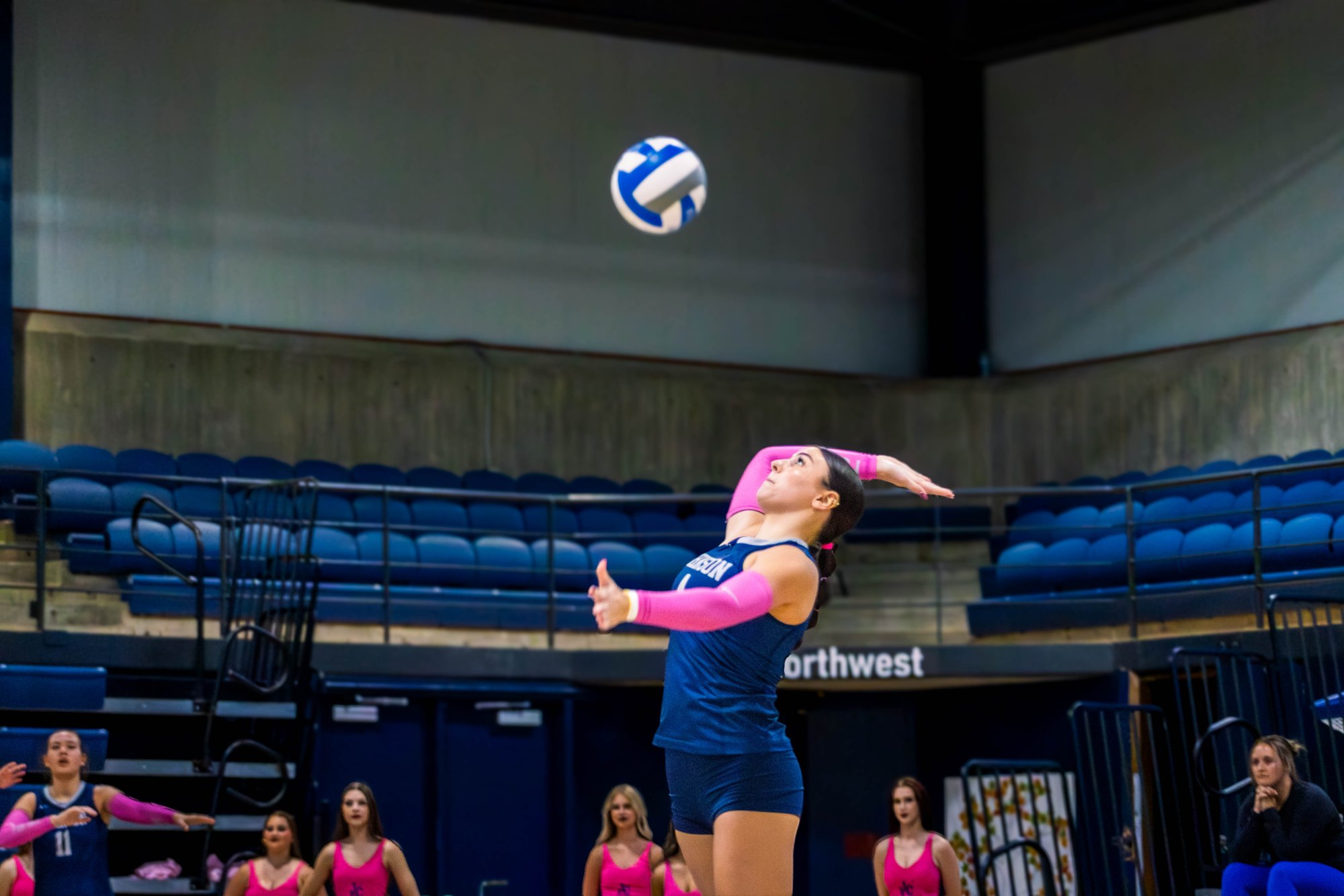 Female volleyball player in navy uniform jumping to strike a ball in an indoor gymnasium.
