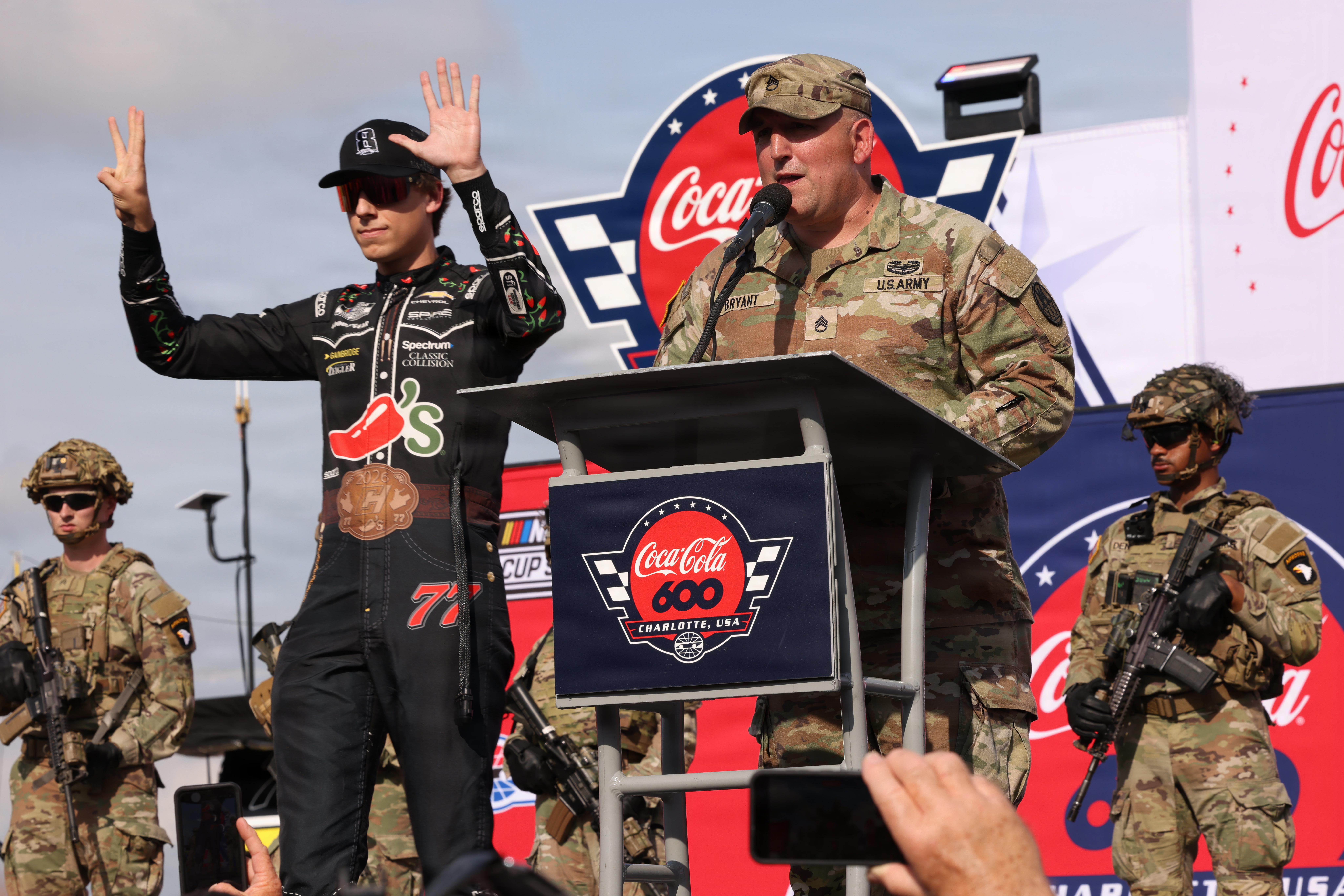 Soldier at a podium with Coca-Cola 600 logo beside a NASCAR driver in a racing suit.